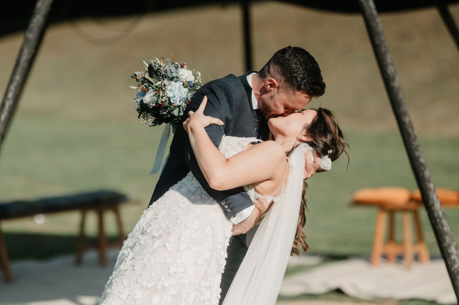 Bride and groom share a romantic kiss during their outdoor wedding ceremony on a sunny day.