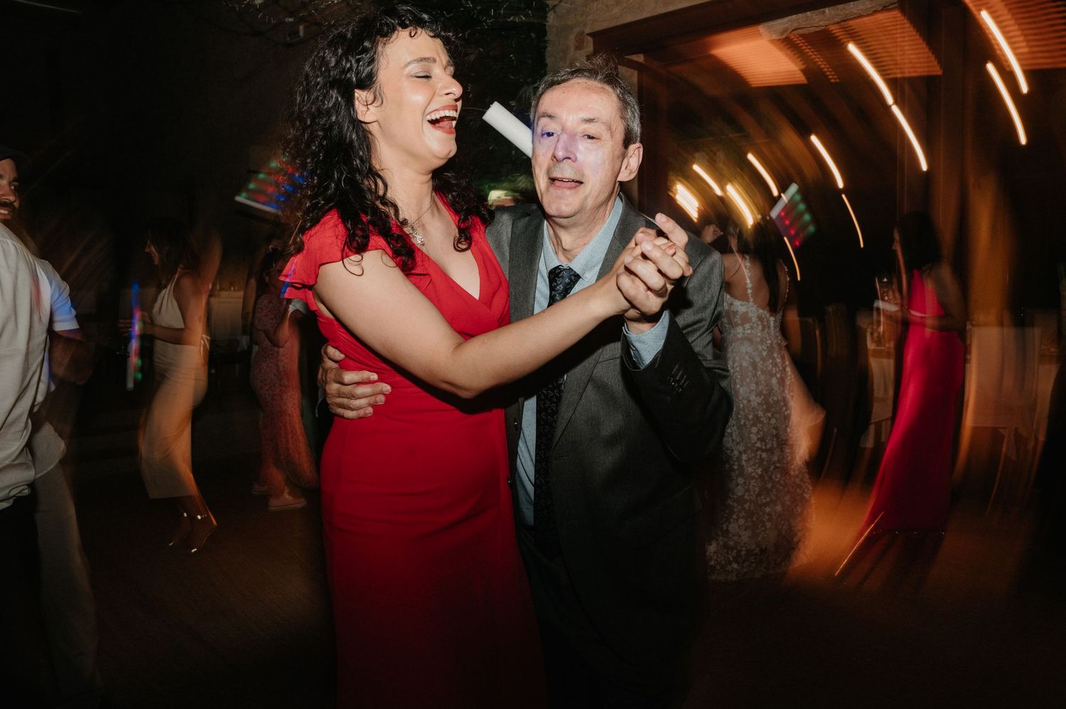 A couple dances together in formal attire at an evening event, with the woman wearing a vibrant red dress.