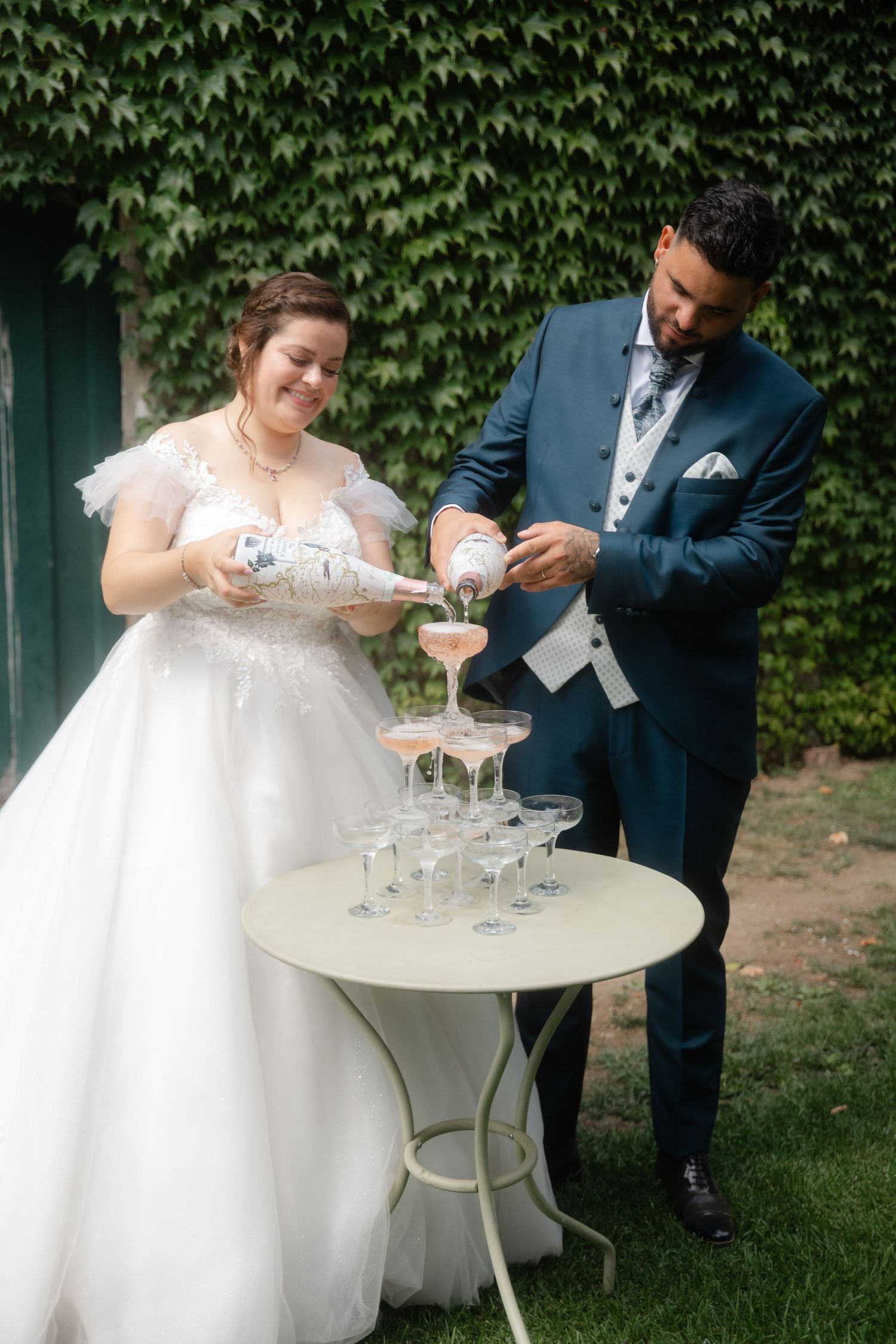 A couple in wedding attire shares drinks at an outdoor garden table with ivy-covered walls in the background.