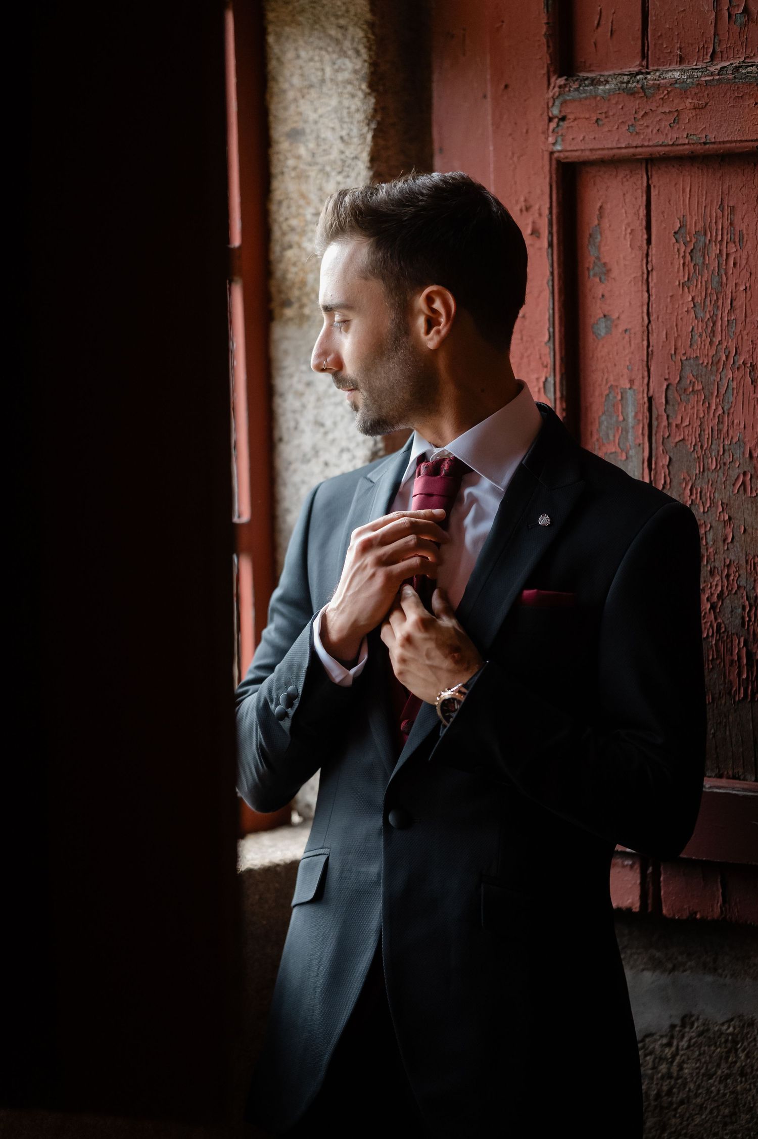 A person in a dark suit adjusts their red tie while standing near a window in moody lighting.