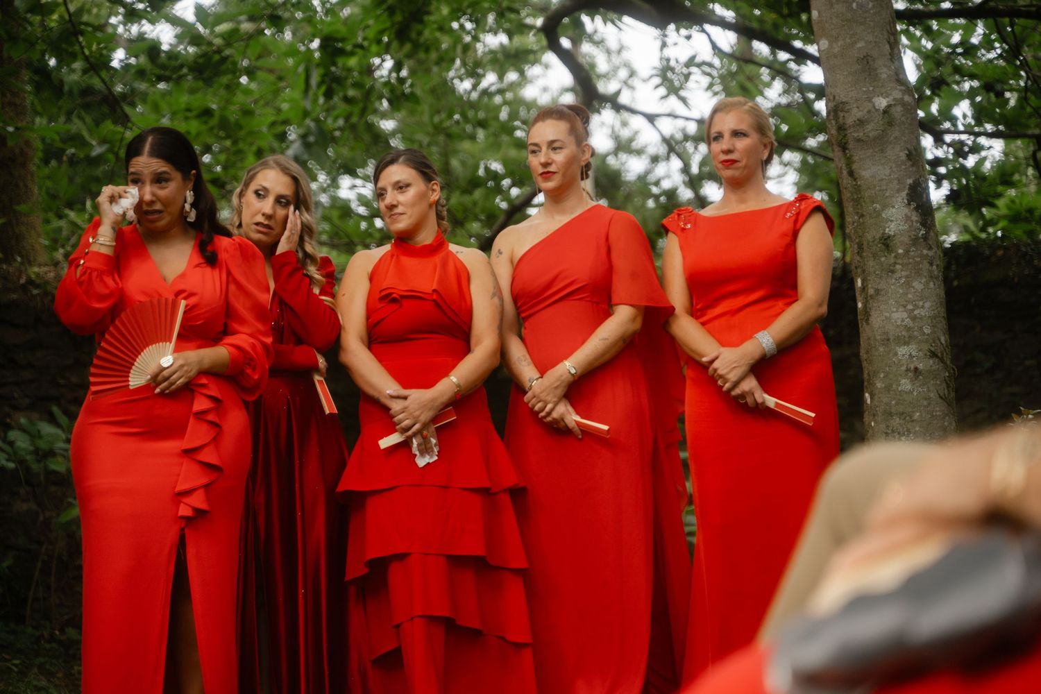 Bridesmaids in matching red dresses pose together outdoors under trees at a wedding celebration.