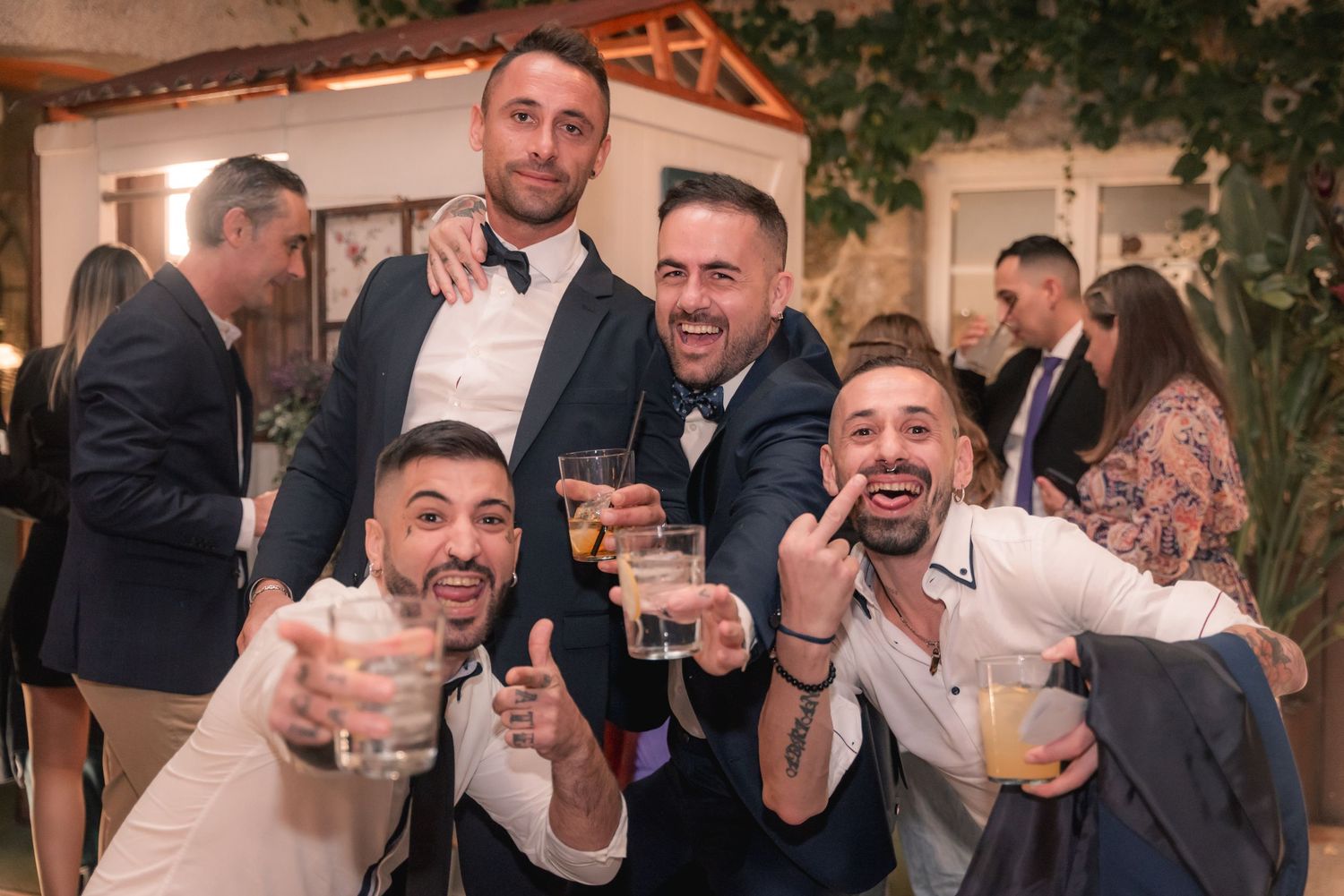 A group of friends raise their drinks in celebration at an indoor event with rustic decor and greenery.