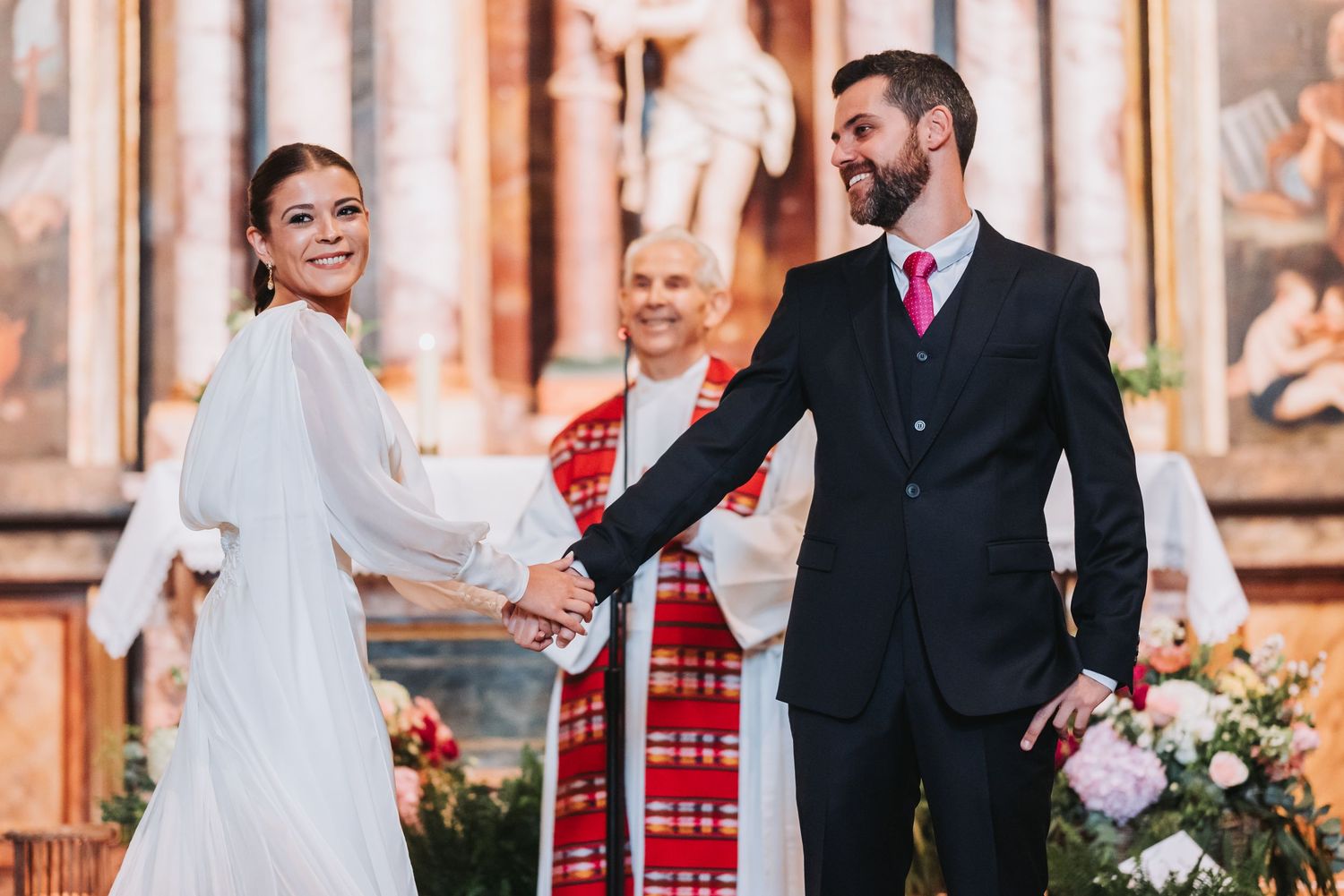 A couple holds hands during their wedding ceremony in a church with ornate decor and pink floral arrangements.