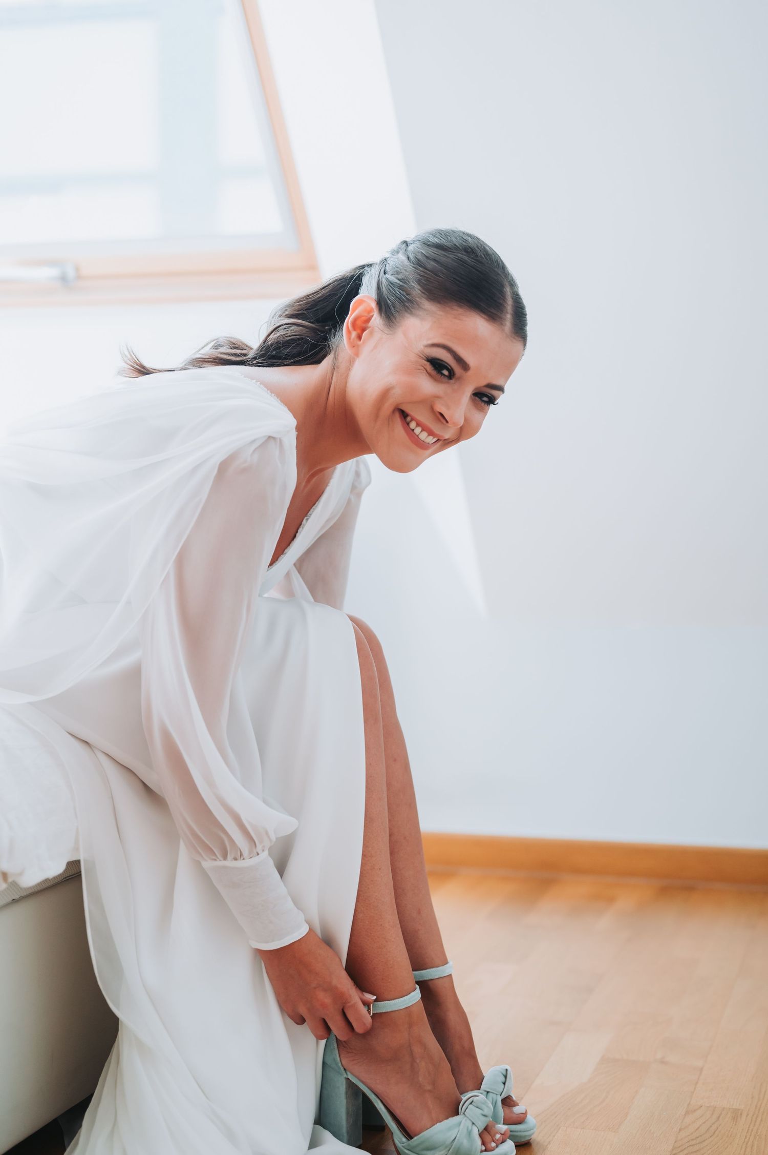 A person in a flowing white wedding dress adjusts their shoe while sitting in a bright room.