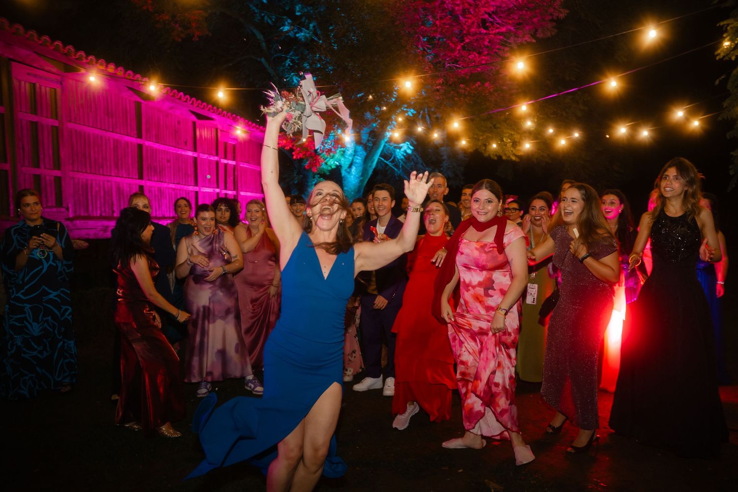 Wedding guests enjoying the dance floor under string lights with vibrant pink and blue ambient lighting.