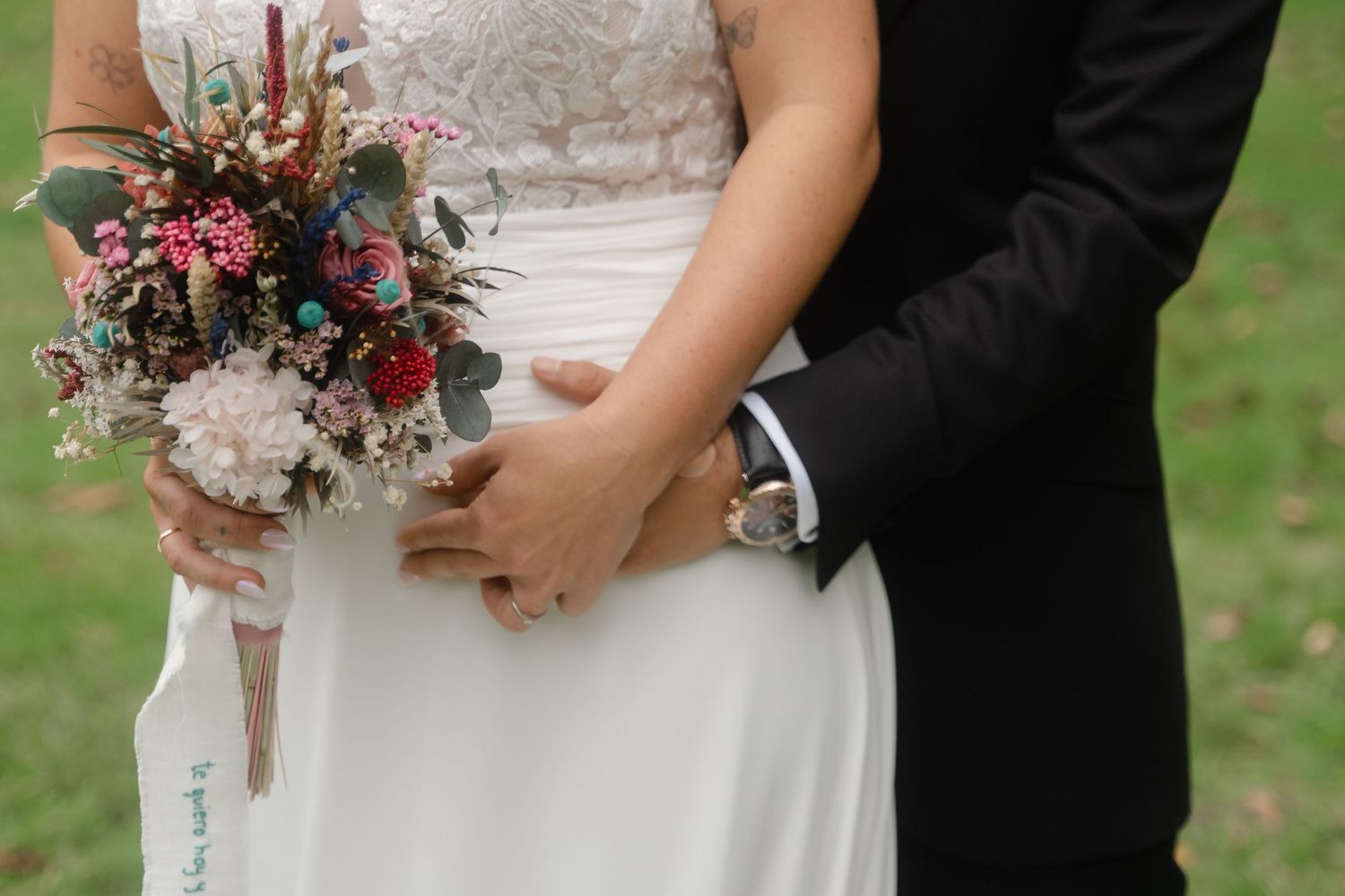 Close-up view of a bridal bouquet with burgundy and blush flowers held against a white wedding dress outdoors.