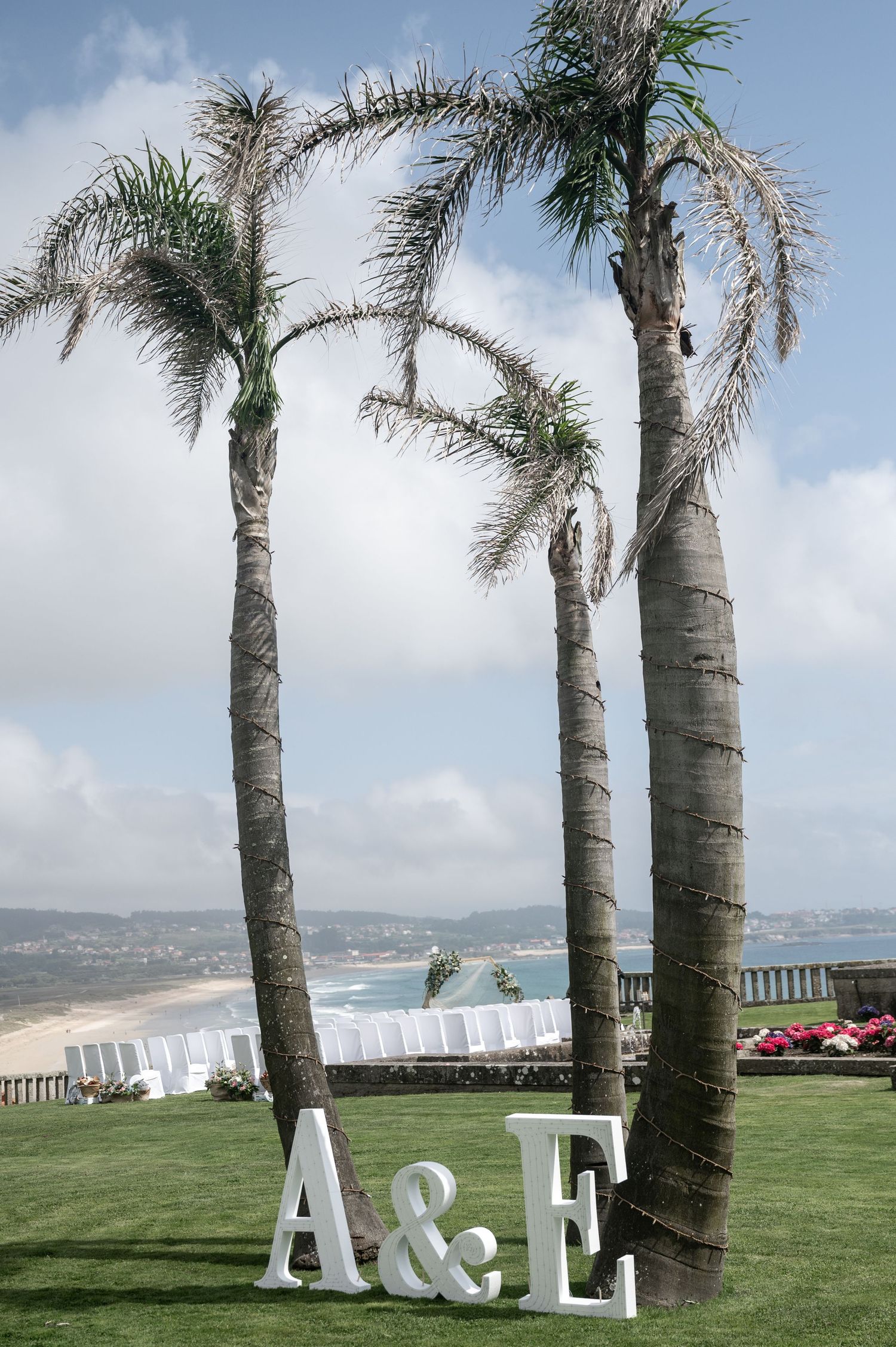 Palm trees line a grassy oceanfront lawn with white chairs and large decorative letters overlooking the coastline.