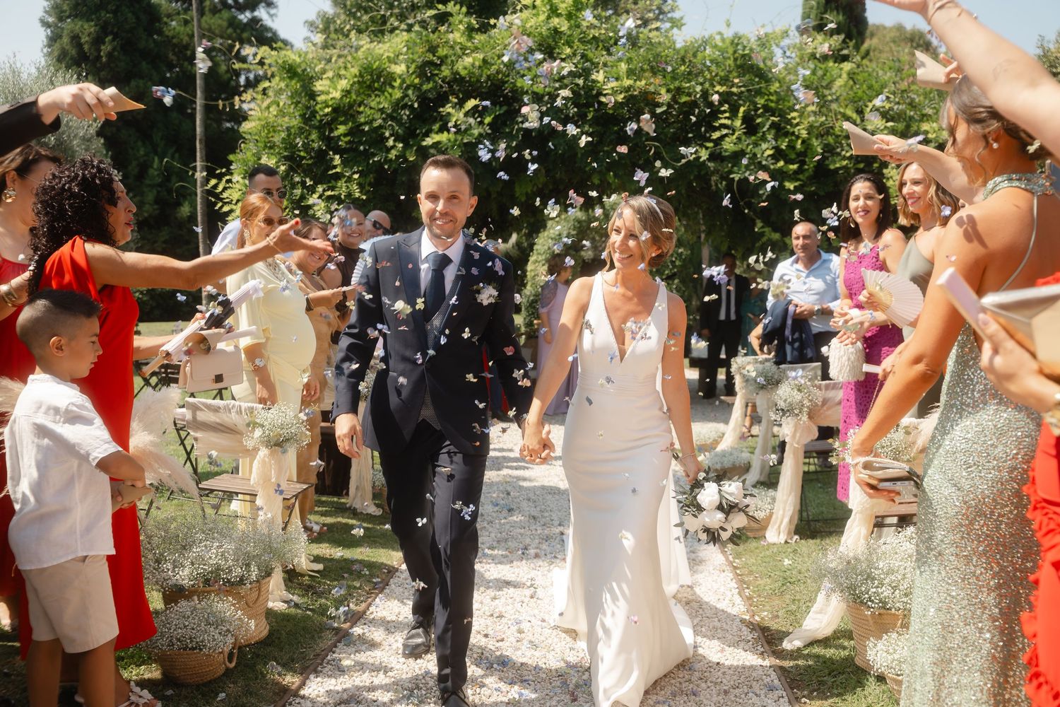 Wedding guests shower newlyweds with confetti along garden path during sunny outdoor celebration.
