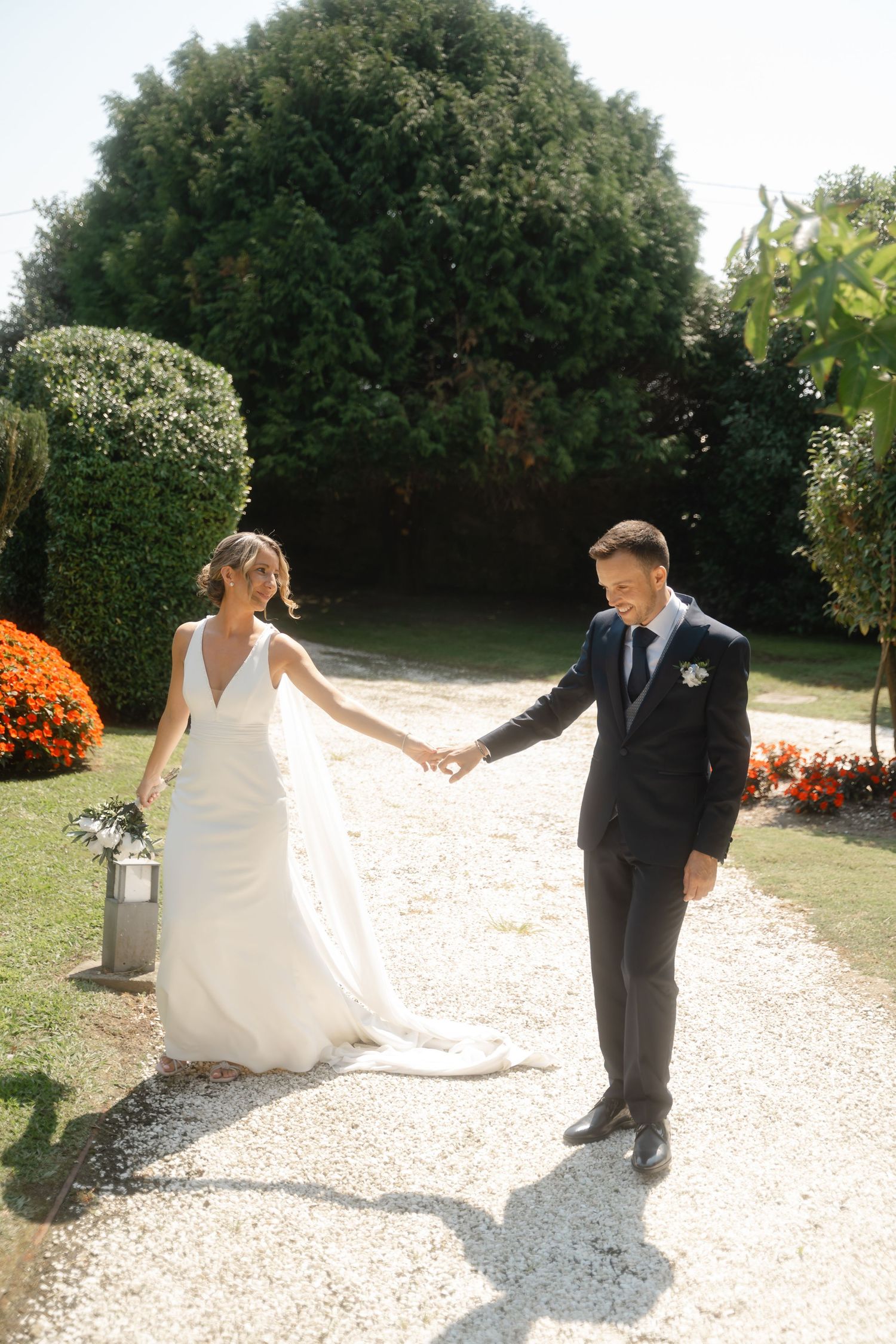 A couple in formal wedding attire poses together on a gravel path surrounded by lush green foliage on a sunny day.