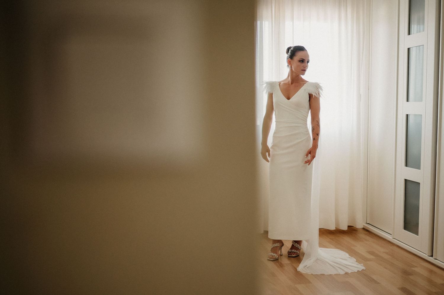 A bride in an elegant white wedding dress stands in a bright hallway with natural light streaming through windows.