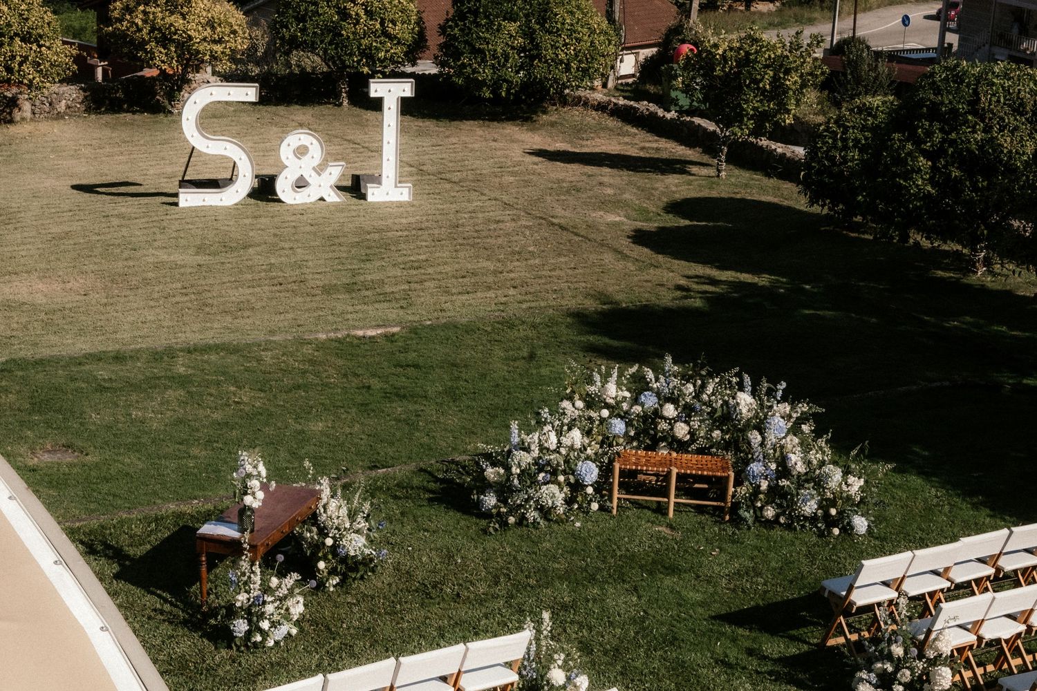 Aerial view of an outdoor wedding ceremony setup with white chairs and floral arrangements in a grassy field.