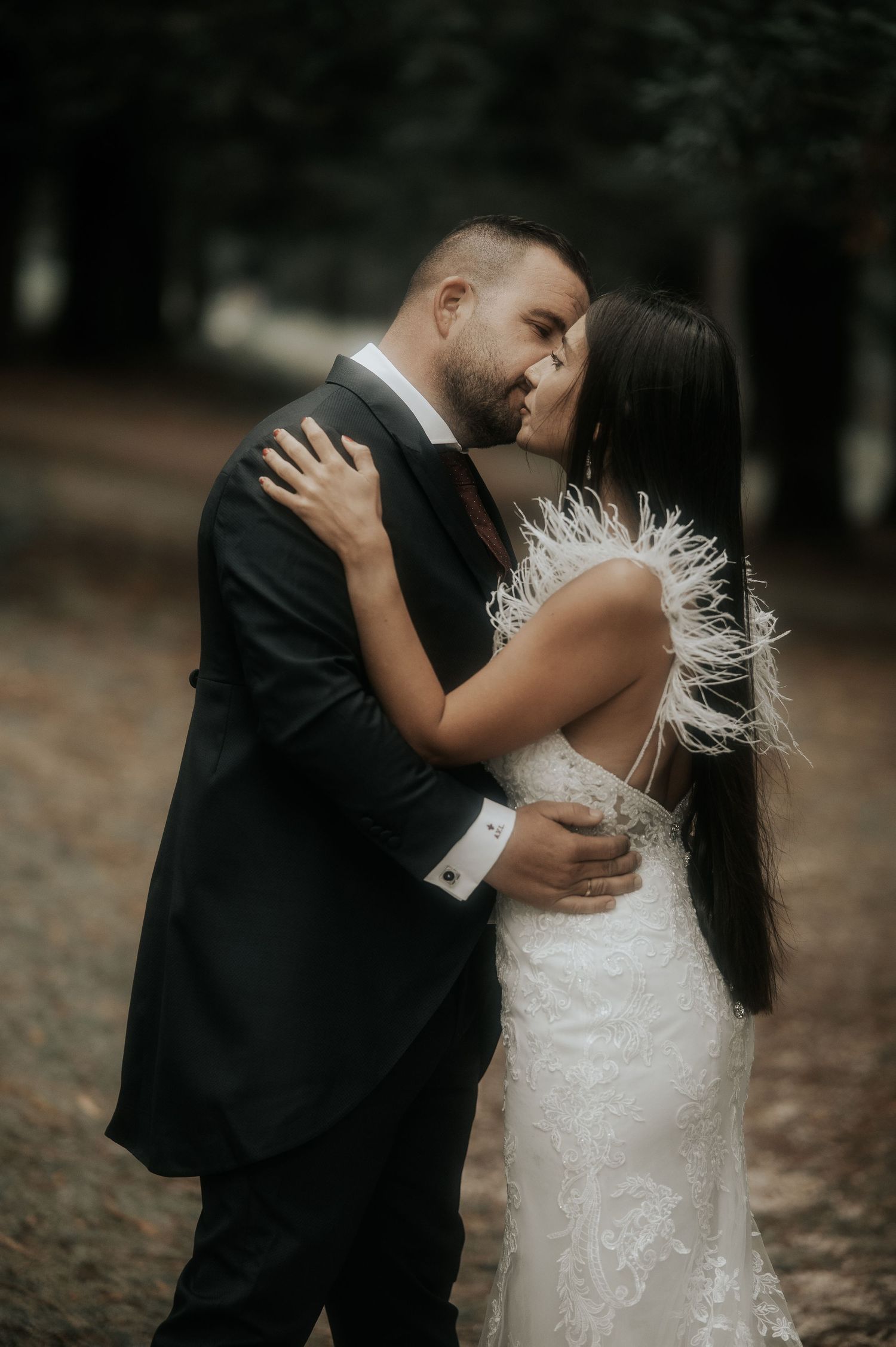 Couple in wedding attire embrace outdoors in moody forest setting with bride wearing feathered white gown.