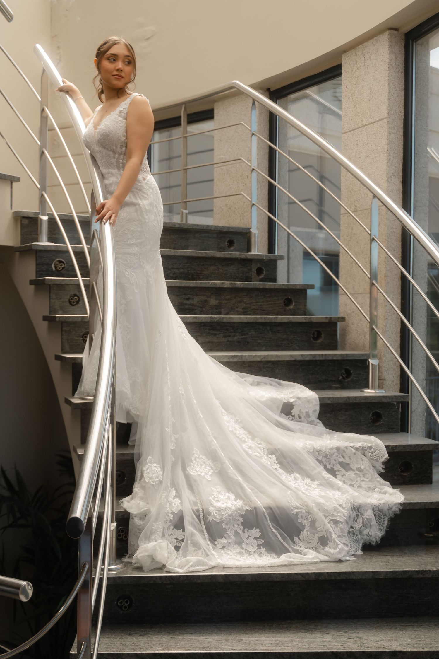 A bride in a flowing lace wedding gown poses elegantly on a modern staircase with metal railings.