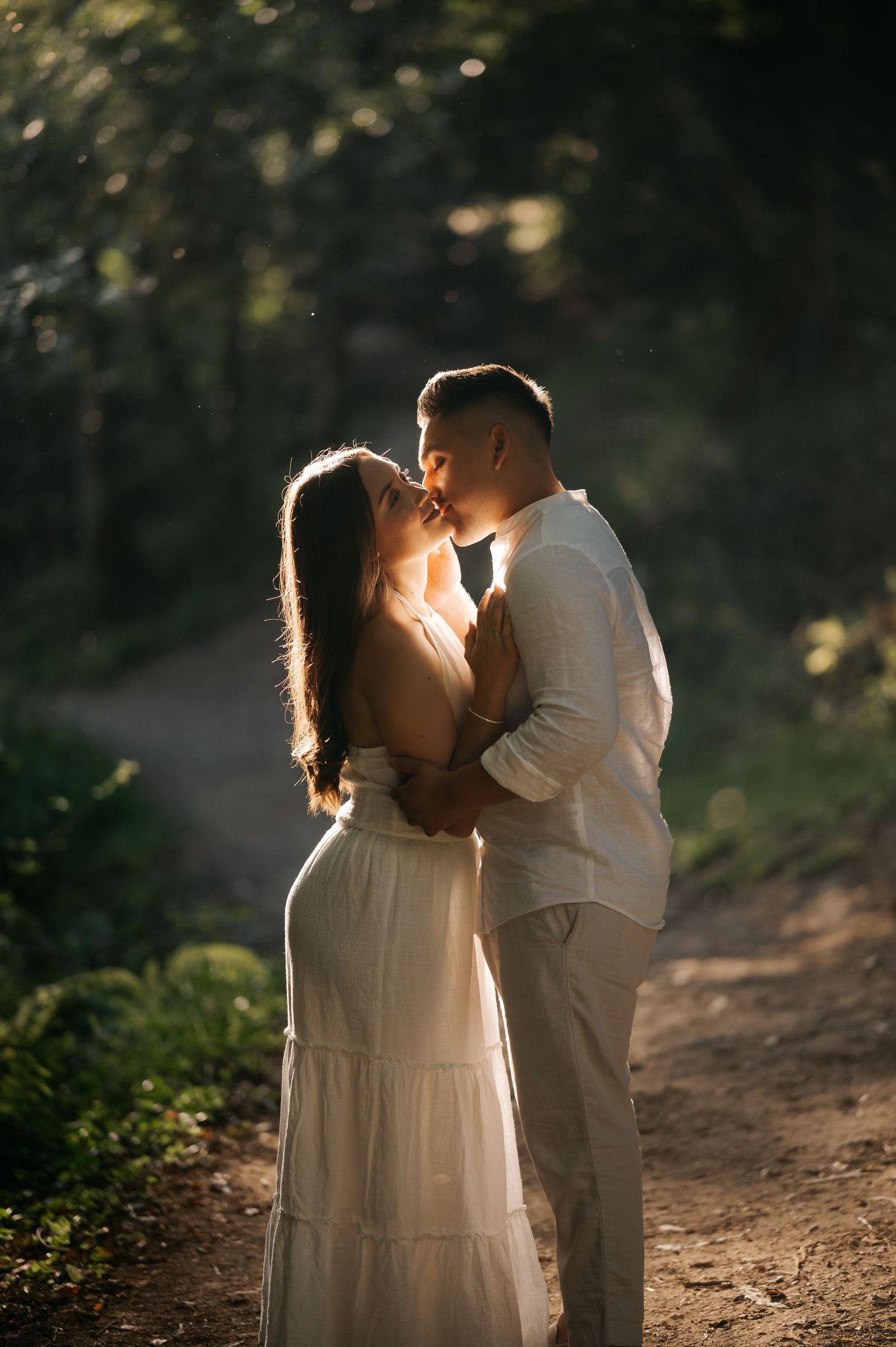 A romantic couple embraces on a sunlit forest path during a golden hour photoshoot.