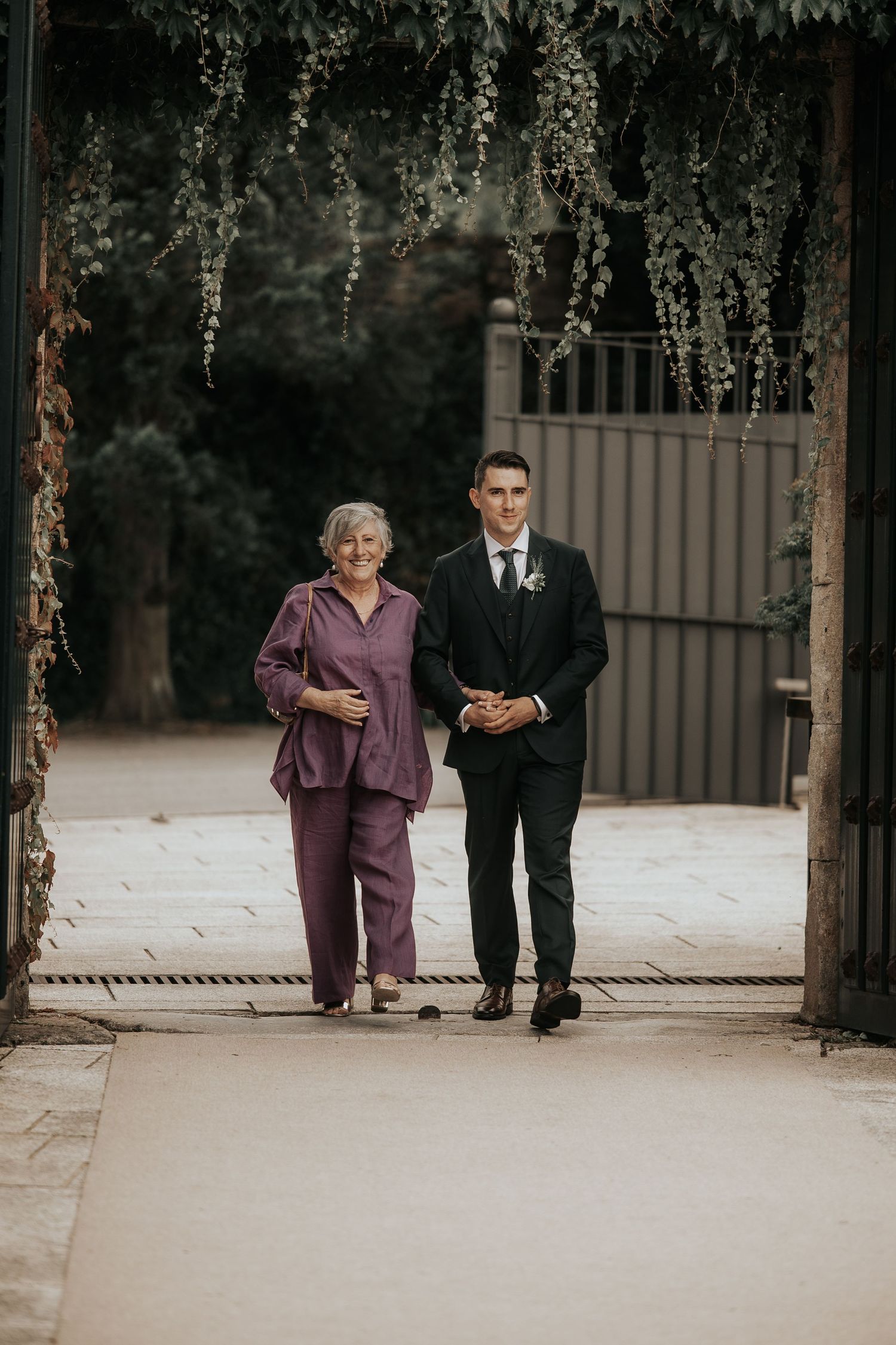 Two people in formal attire walk together on a path surrounded by trees.