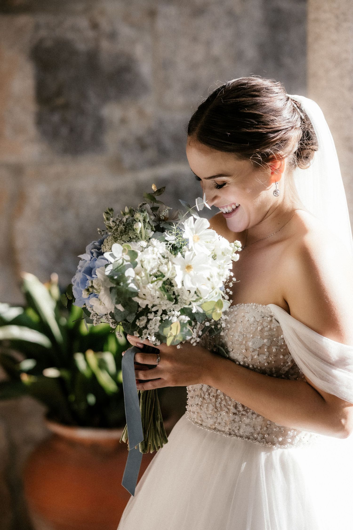 A bride in a strapless beaded gown smiles while holding a white and blue floral bouquet with trailing ribbons.