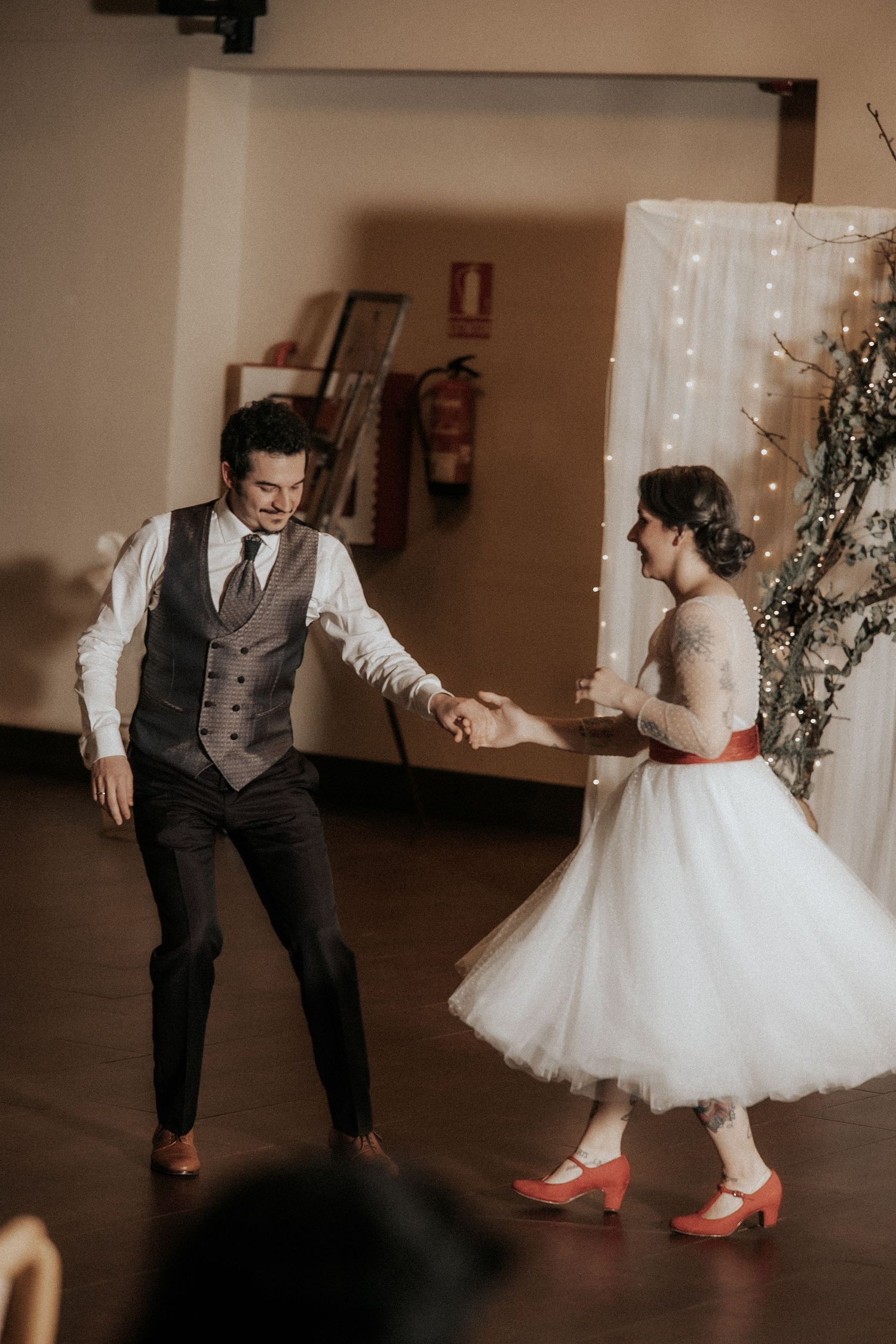 A couple dances playfully at a wedding reception, with the bride in a short white dress and red heels.
