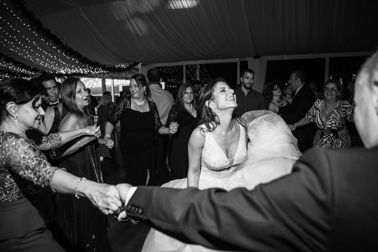 Wedding guests dance and celebrate under twinkling lights at an evening reception.