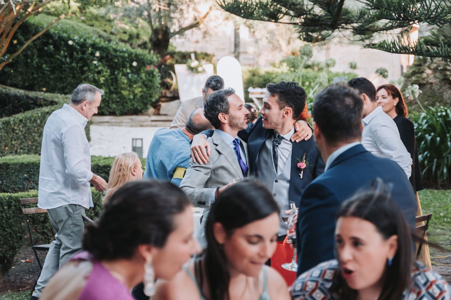 A group of people gathered outdoors for a casual social event with tables and chairs in a garden setting.