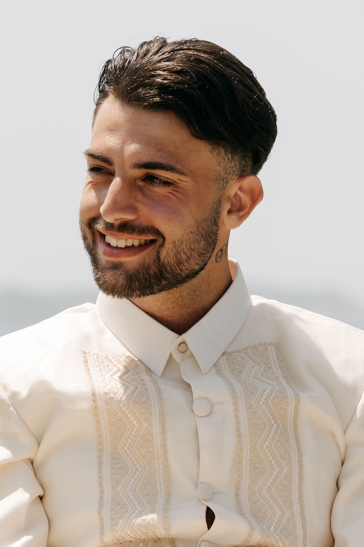 A person wearing a cream colored embroidered shirt smiles warmly against a light background.