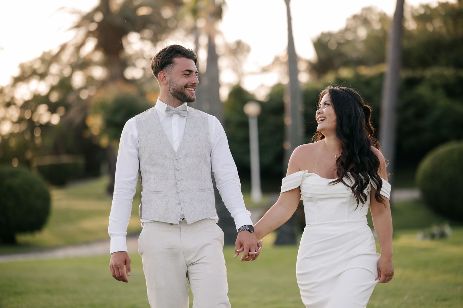 A couple in white wedding attire walks hand in hand through a sunlit garden during golden hour.