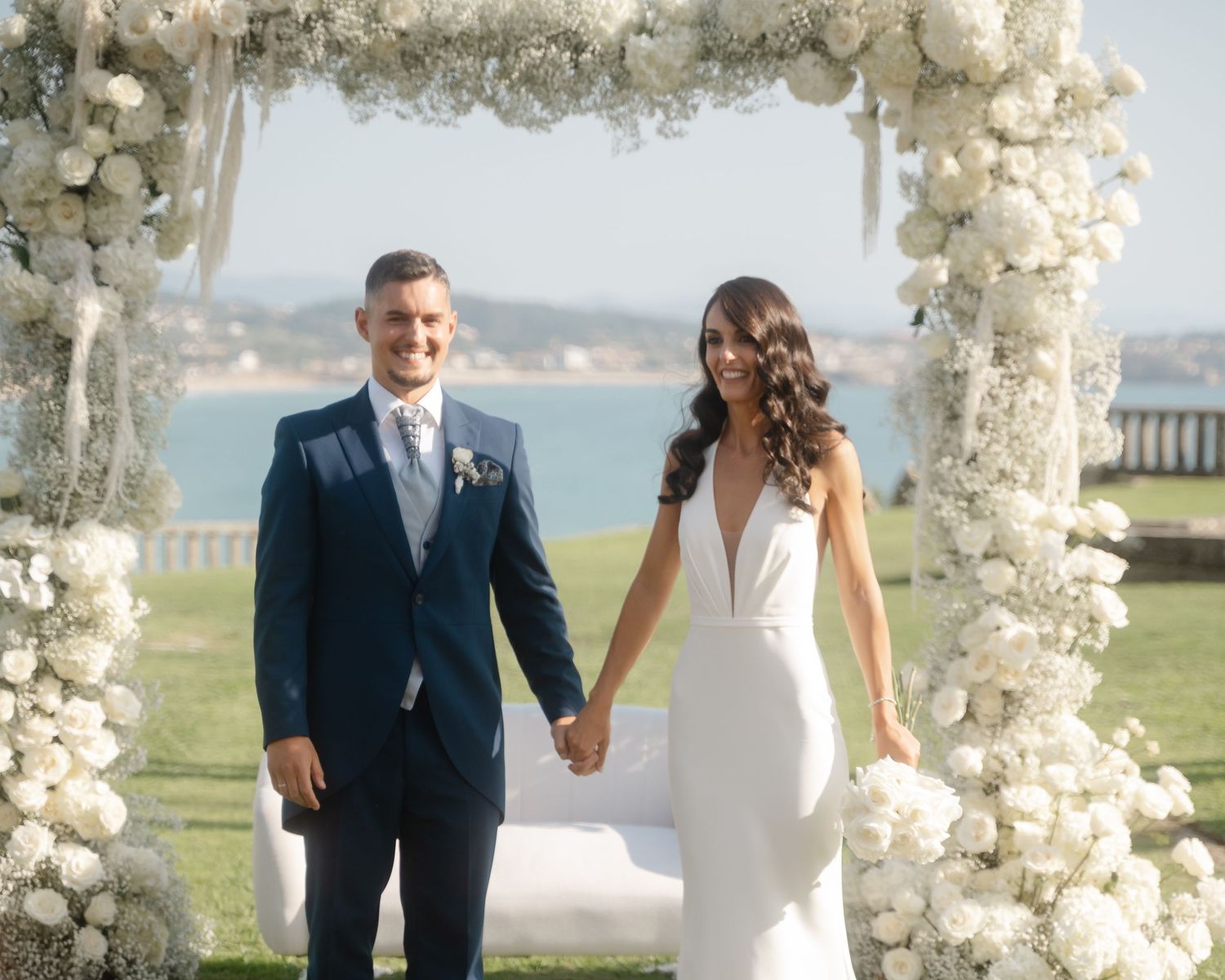 A newlywed couple stands under a white floral arch overlooking a scenic coastal view on their wedding day.