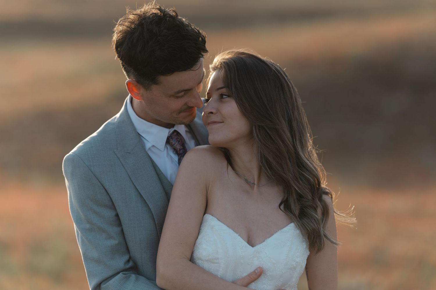 Bride and groom share a tender moment in warm golden sunset light in open field.