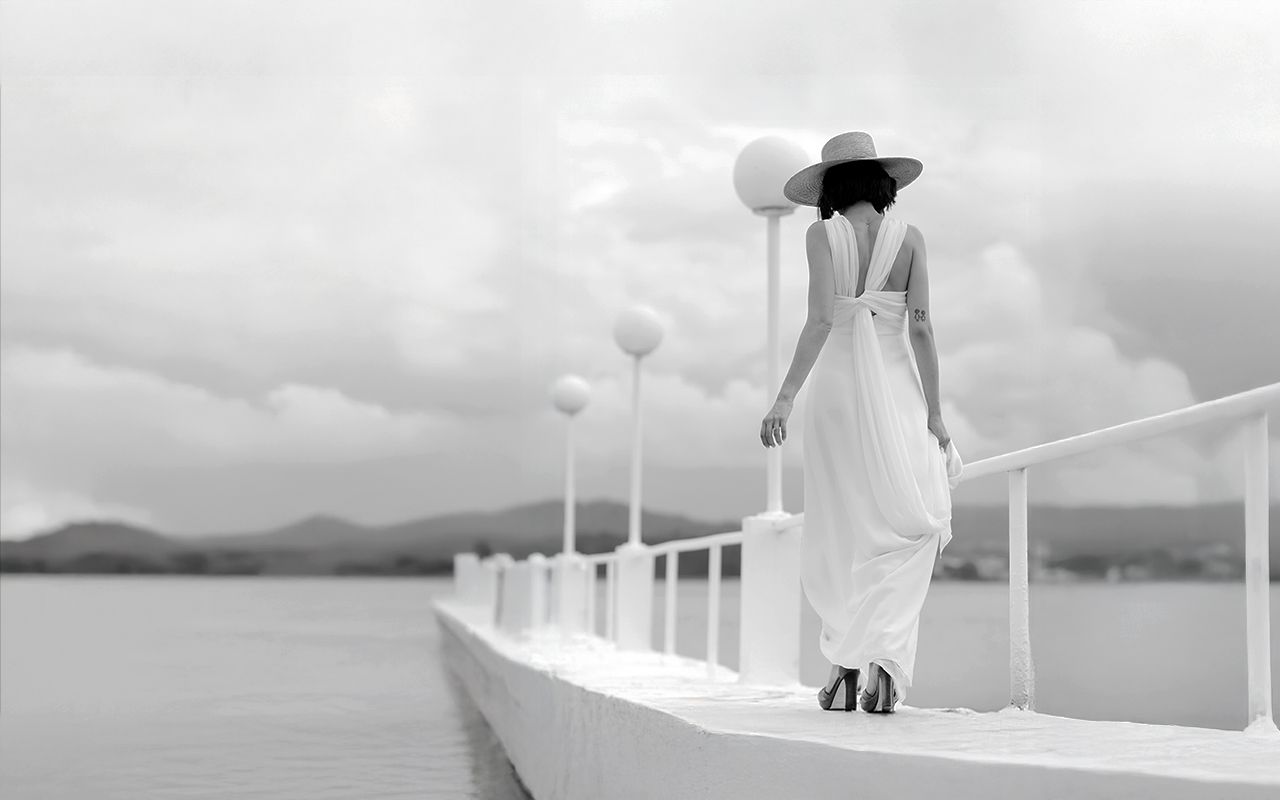 Woman in white dress and hat stands on pier overlooking misty mountains in elegant black and white photo.