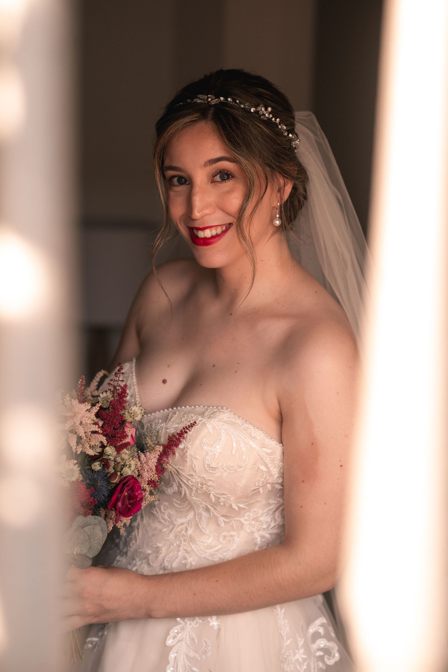 A bridal portrait captured through white columns showing a strapless gown and red floral bouquet.