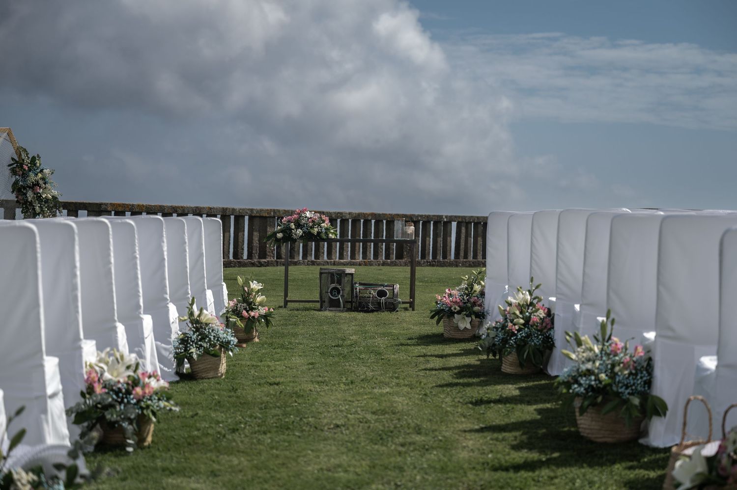White chairs arranged in rows on green grass for an outdoor wedding ceremony with flower decorations and cloudy sky.