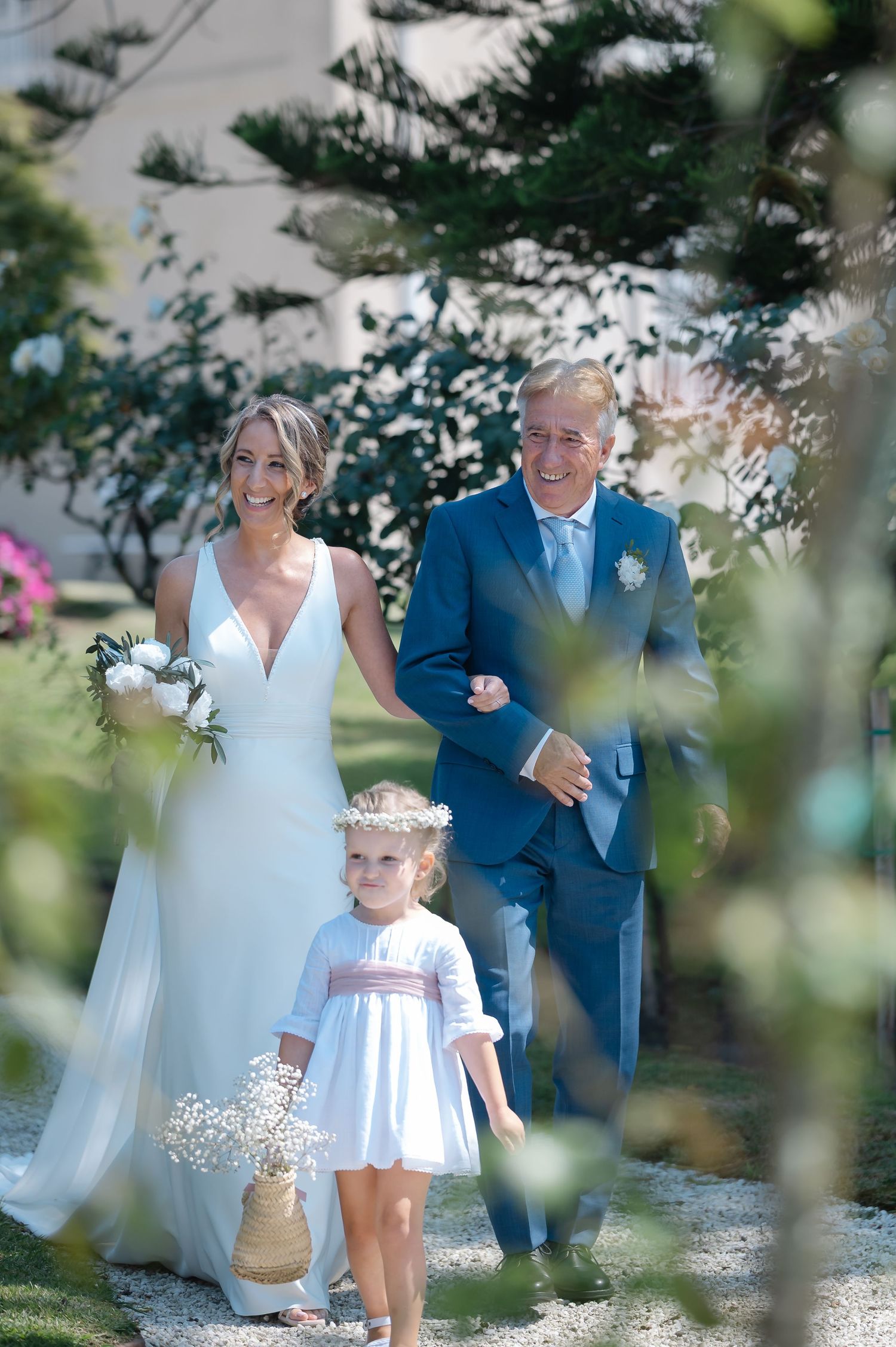 A wedding party walks together outdoors on a sunny day through a garden setting with blurred foliage in the foreground.