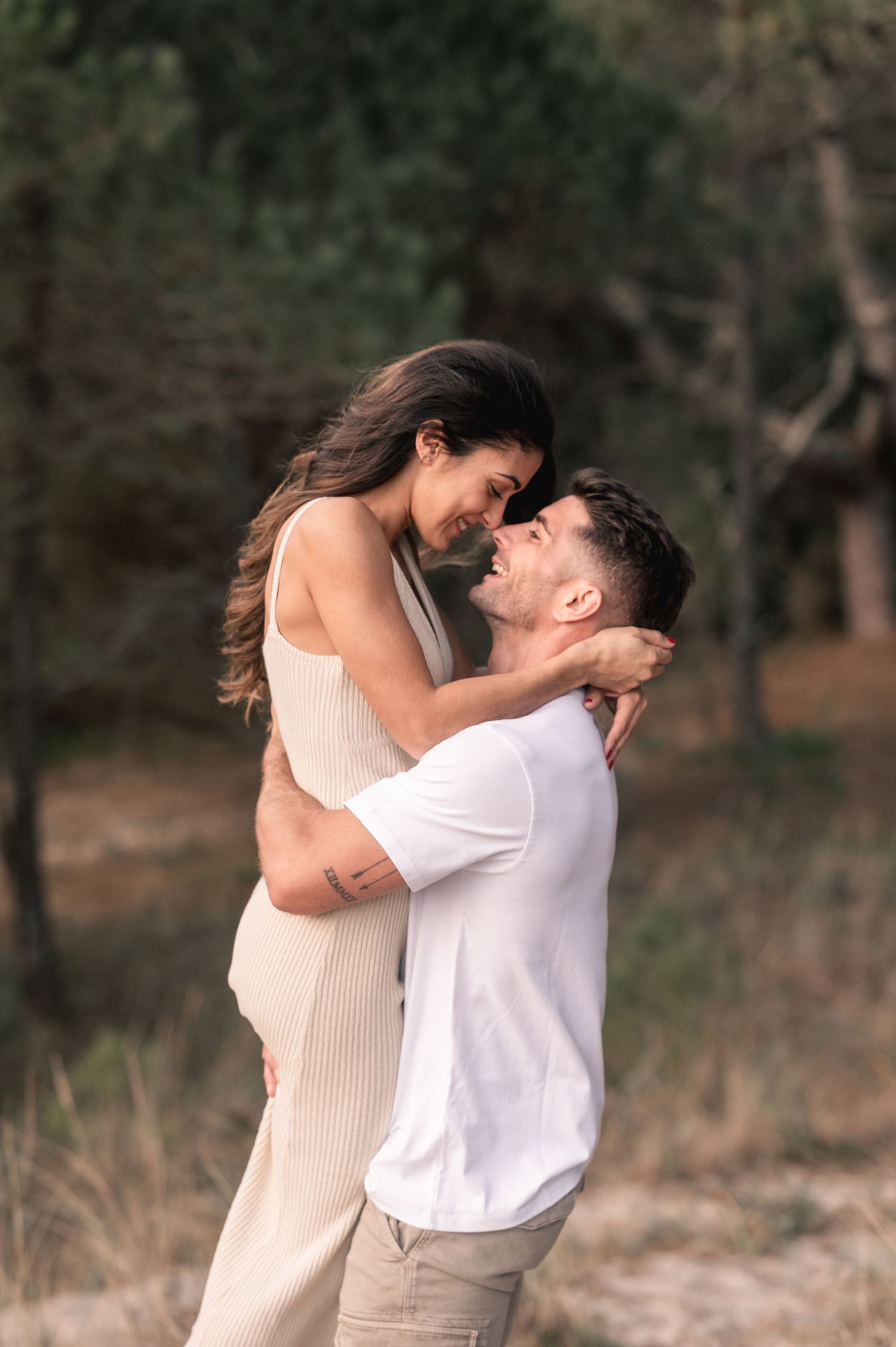 A romantic couple embraces and spins joyfully in a natural outdoor setting with trees in the background.