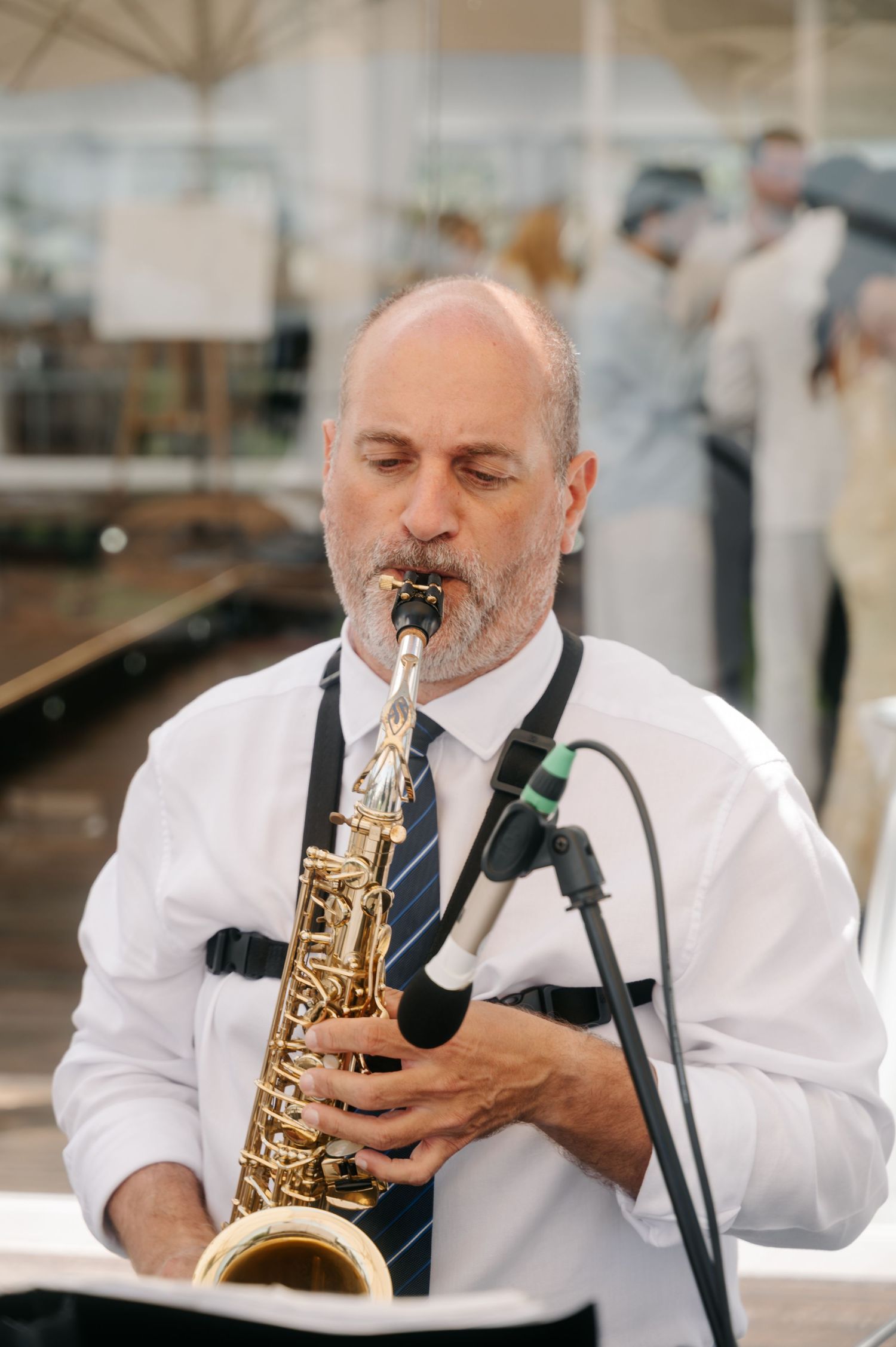 A musician in a white shirt and tie plays saxophone at an outdoor event with a microphone stand visible.