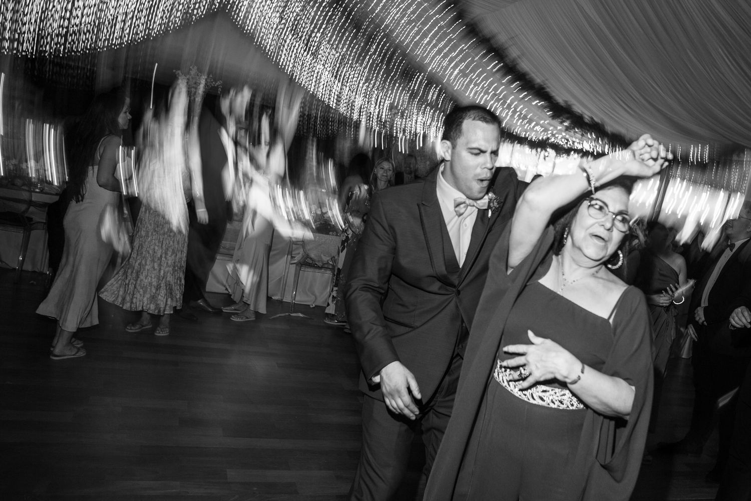 Black and white photo captures joyful dancing at wedding reception under decorative lighting.