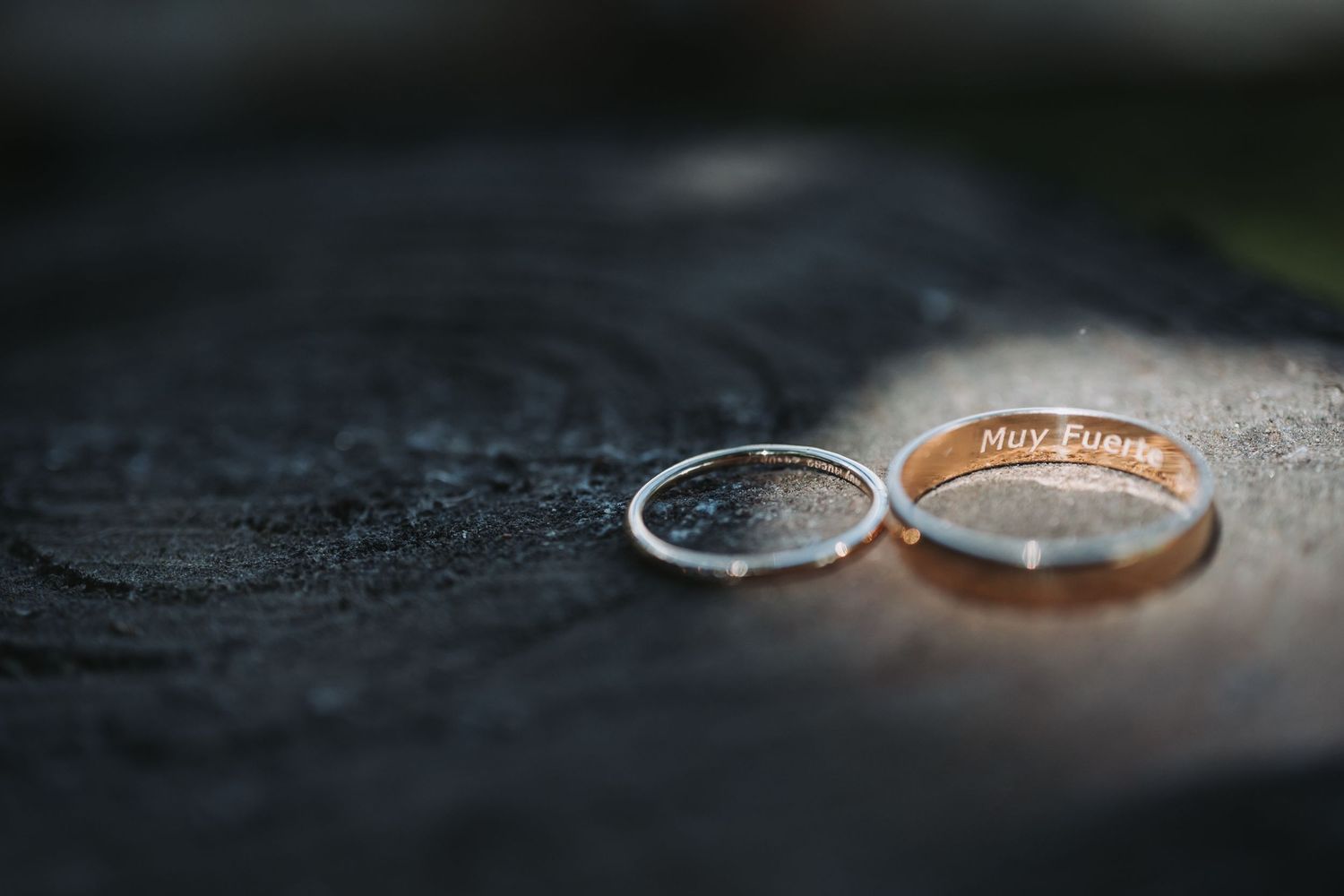 Two wedding rings, one silver and one rose gold, lying on a dark textured surface with soft lighting.