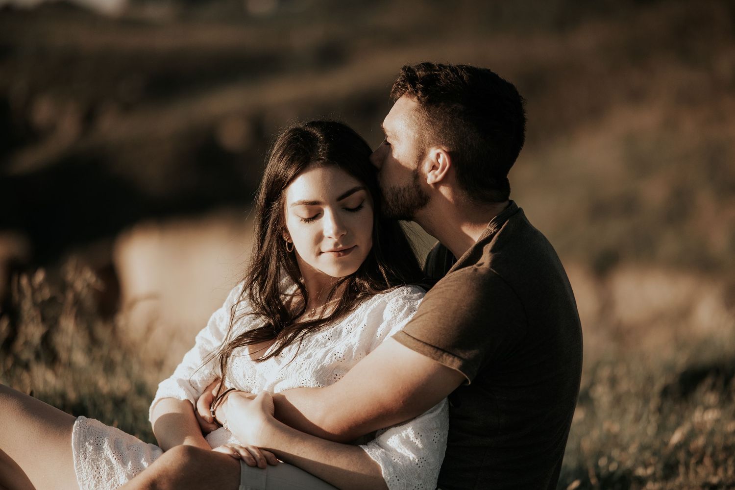 Romantic couple embraces on grassy hillside during golden hour photoshoot in sepia tones.