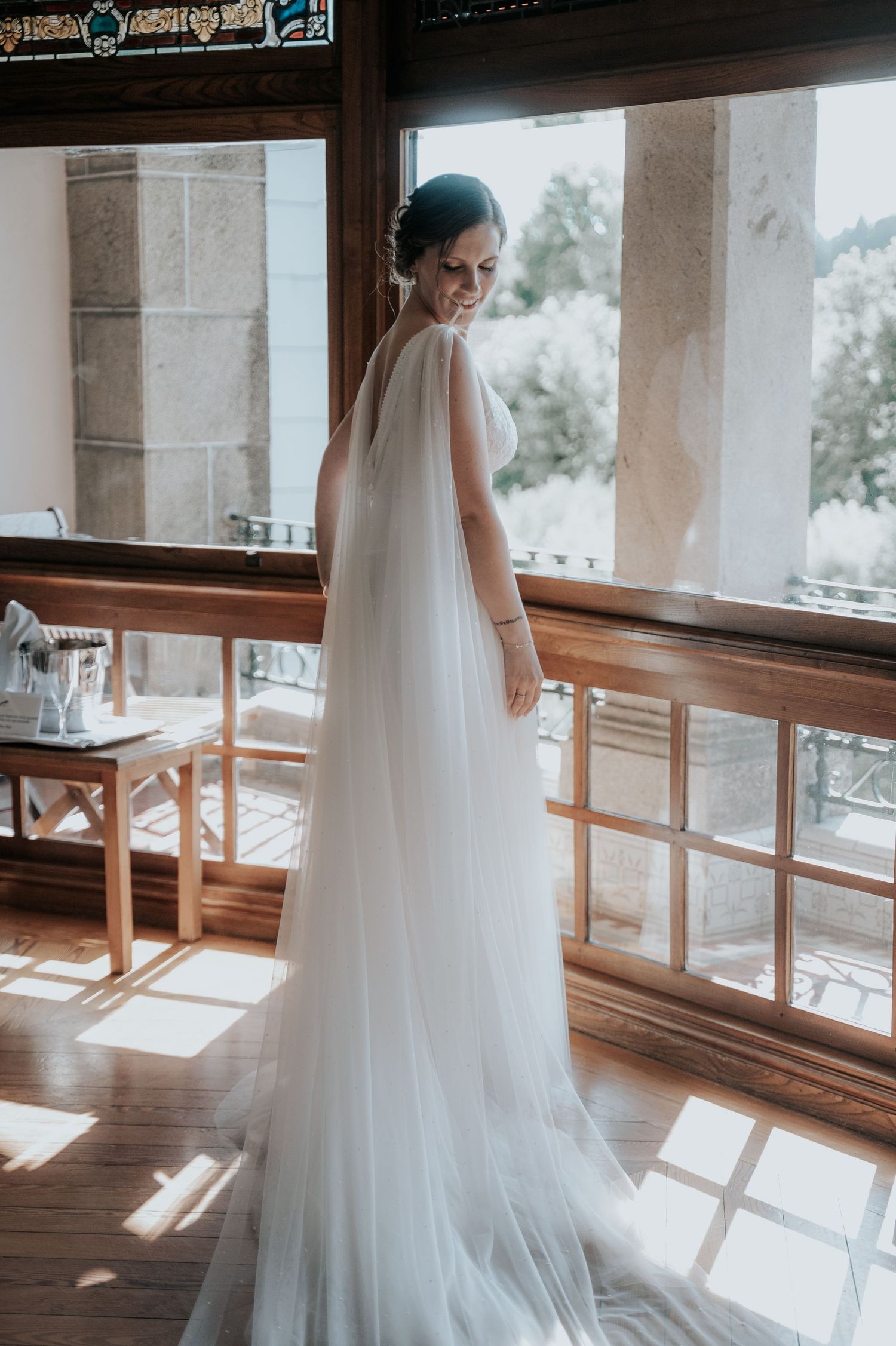 Elegant bride in flowing white gown and veil stands near large windows in sunlit room with wooden floors.