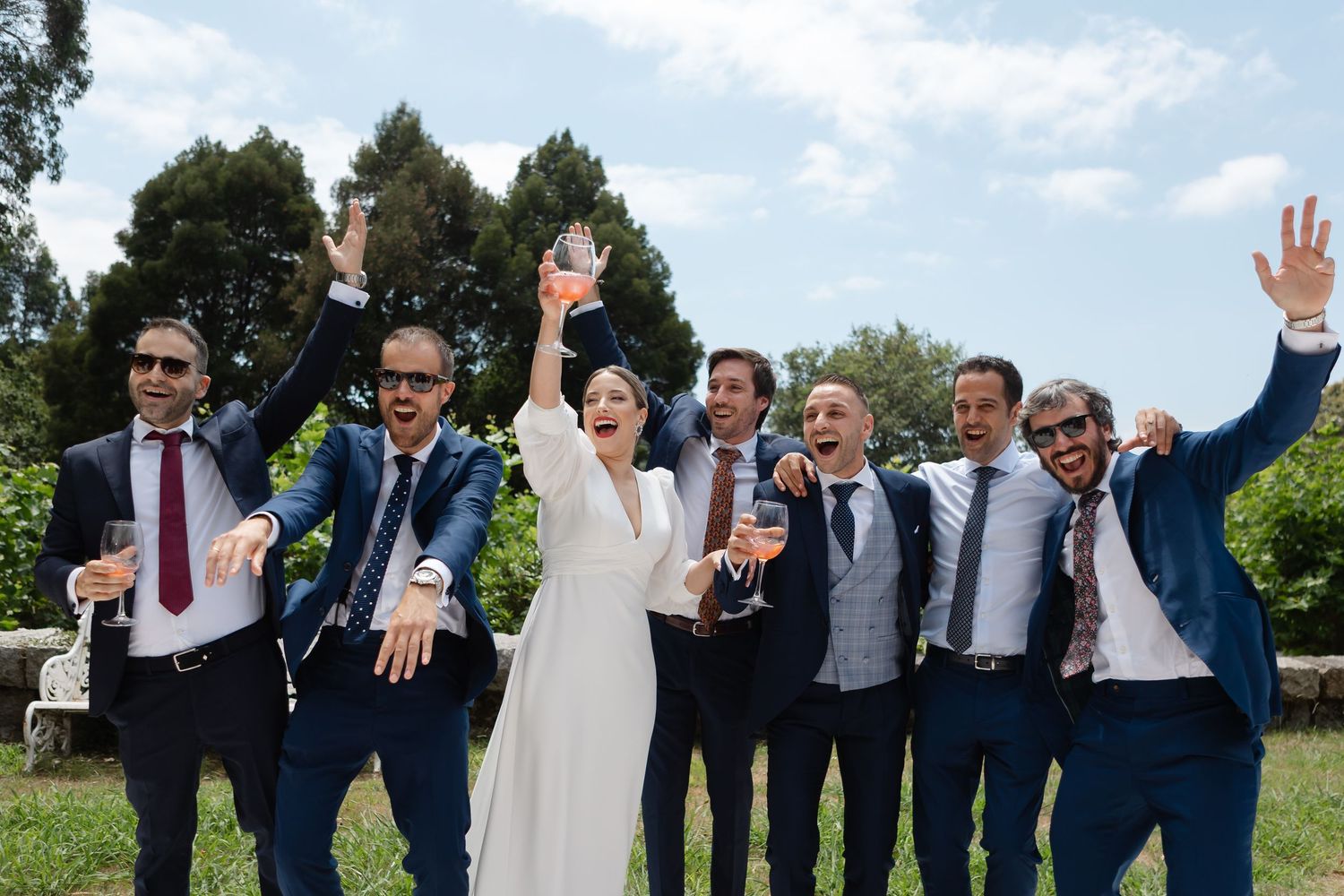 Wedding party members in navy suits and a white dress pose together outdoors on a sunny day.