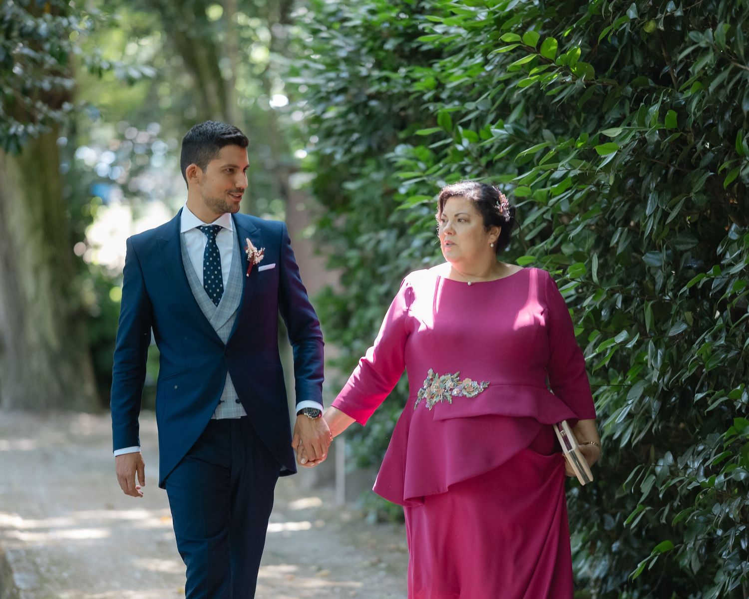 A couple in formal attire walks together through a lush garden path during a wedding photo session.