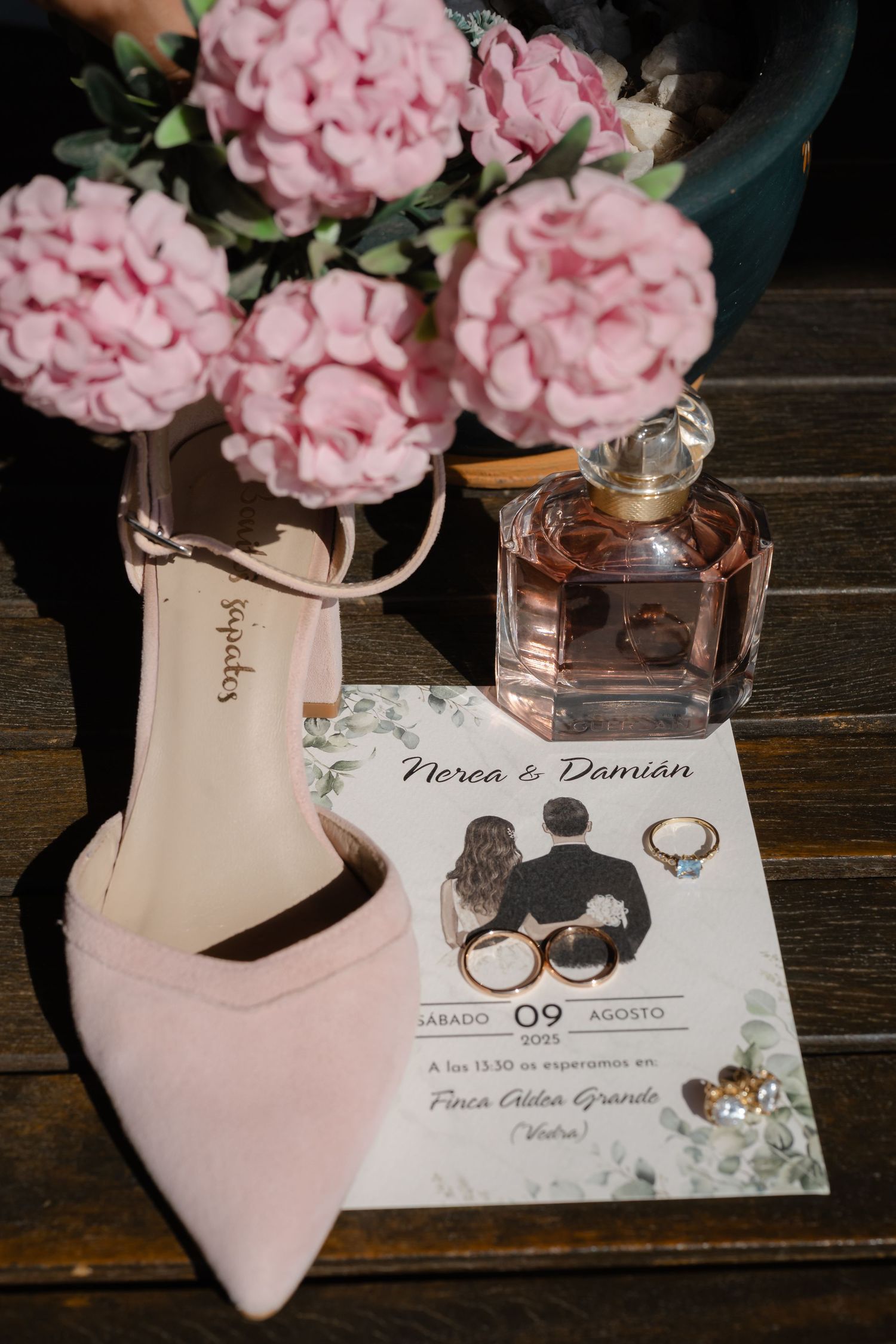 Wedding details featuring pink carnations, high heels, perfume bottle and invitation card on rustic wooden surface.