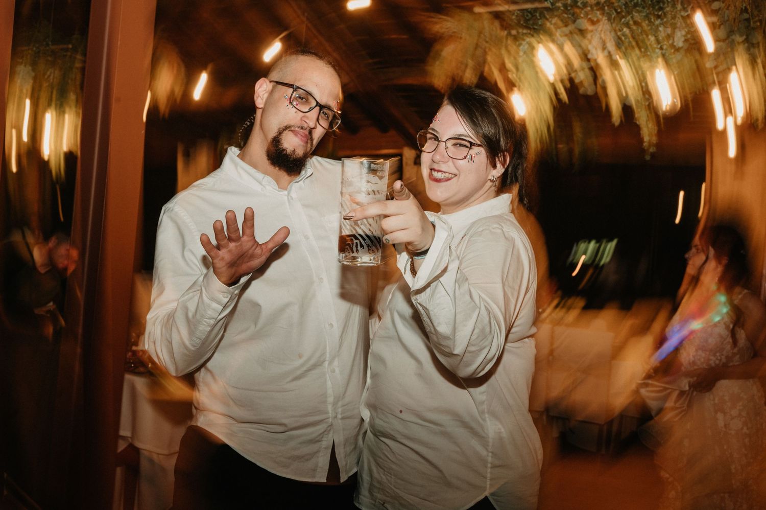 Two guests posing playfully with drinks at an evening celebration under warm lighting.