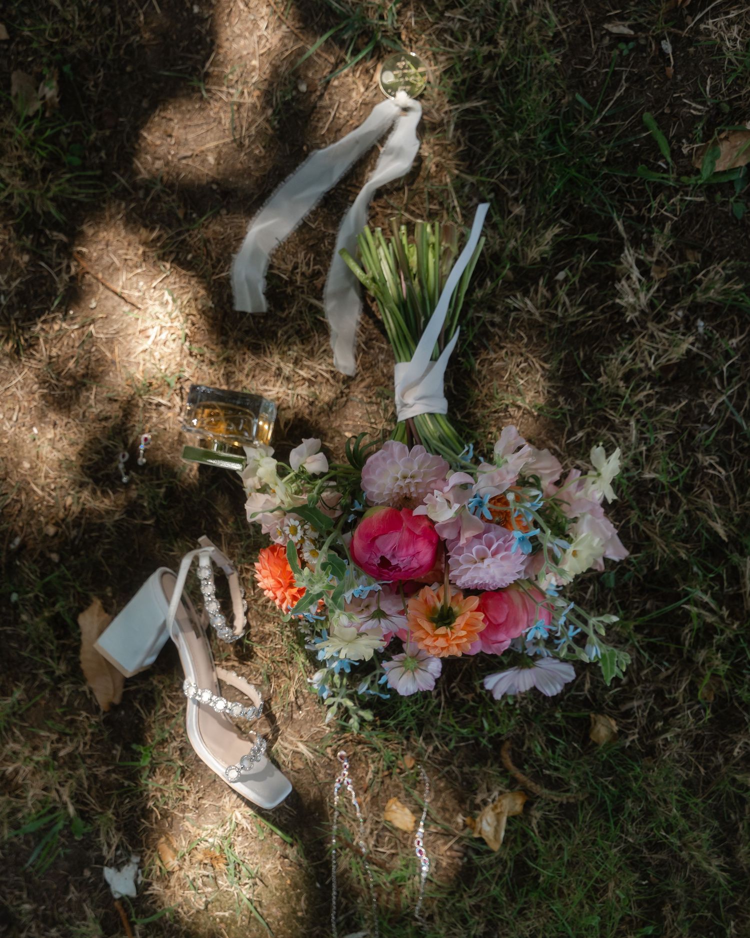White wedding shoes and colorful floral bouquet arranged on natural ground in dappled sunlight.