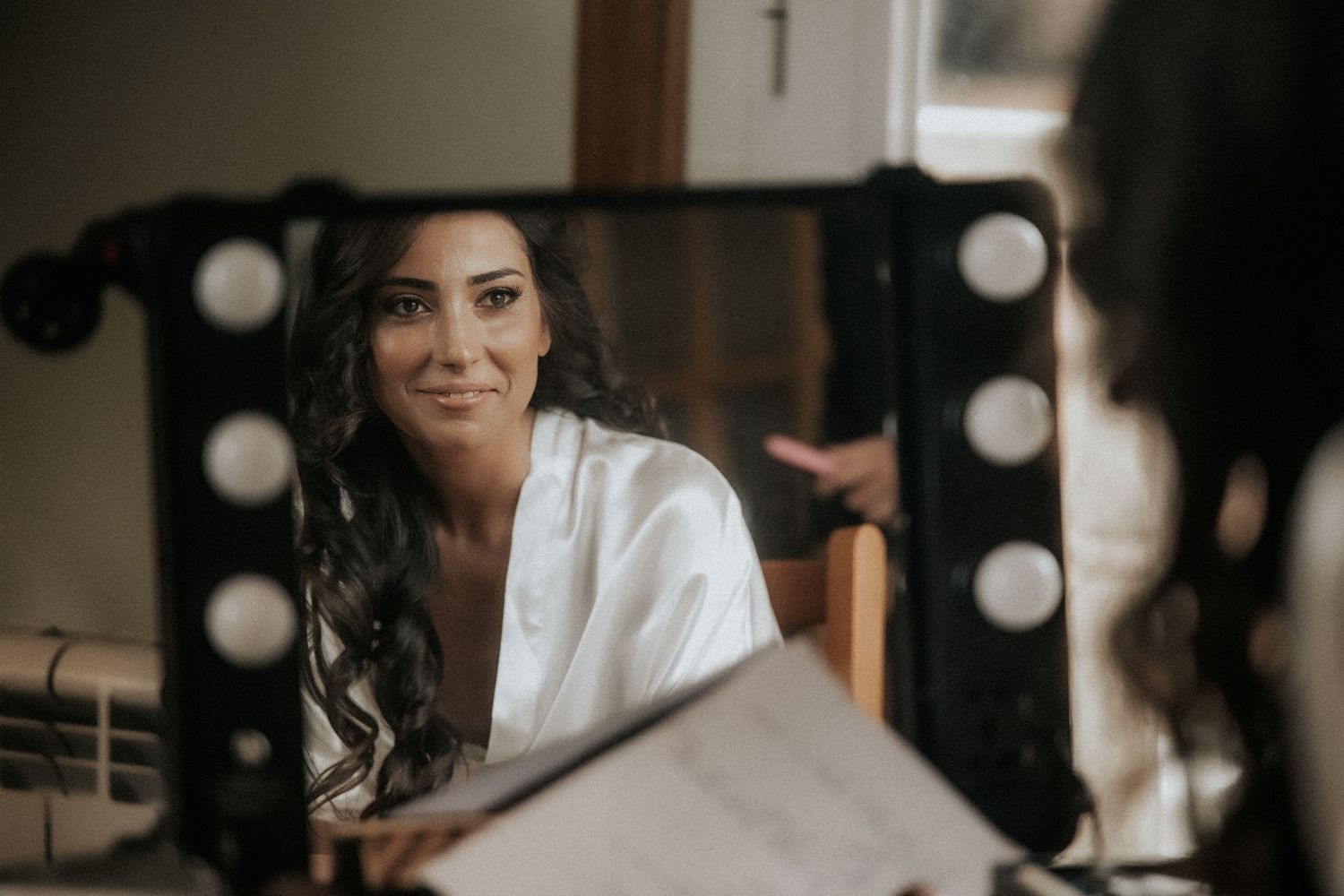A person wearing a white robe sits smiling in front of a mirror with professional lighting and makeup setup.