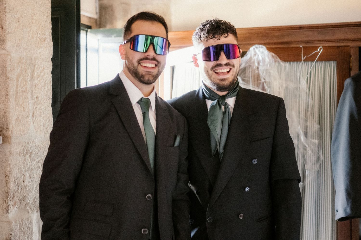 A group of groomsmen in formal attire stand together smiling in a rustic indoor setting.