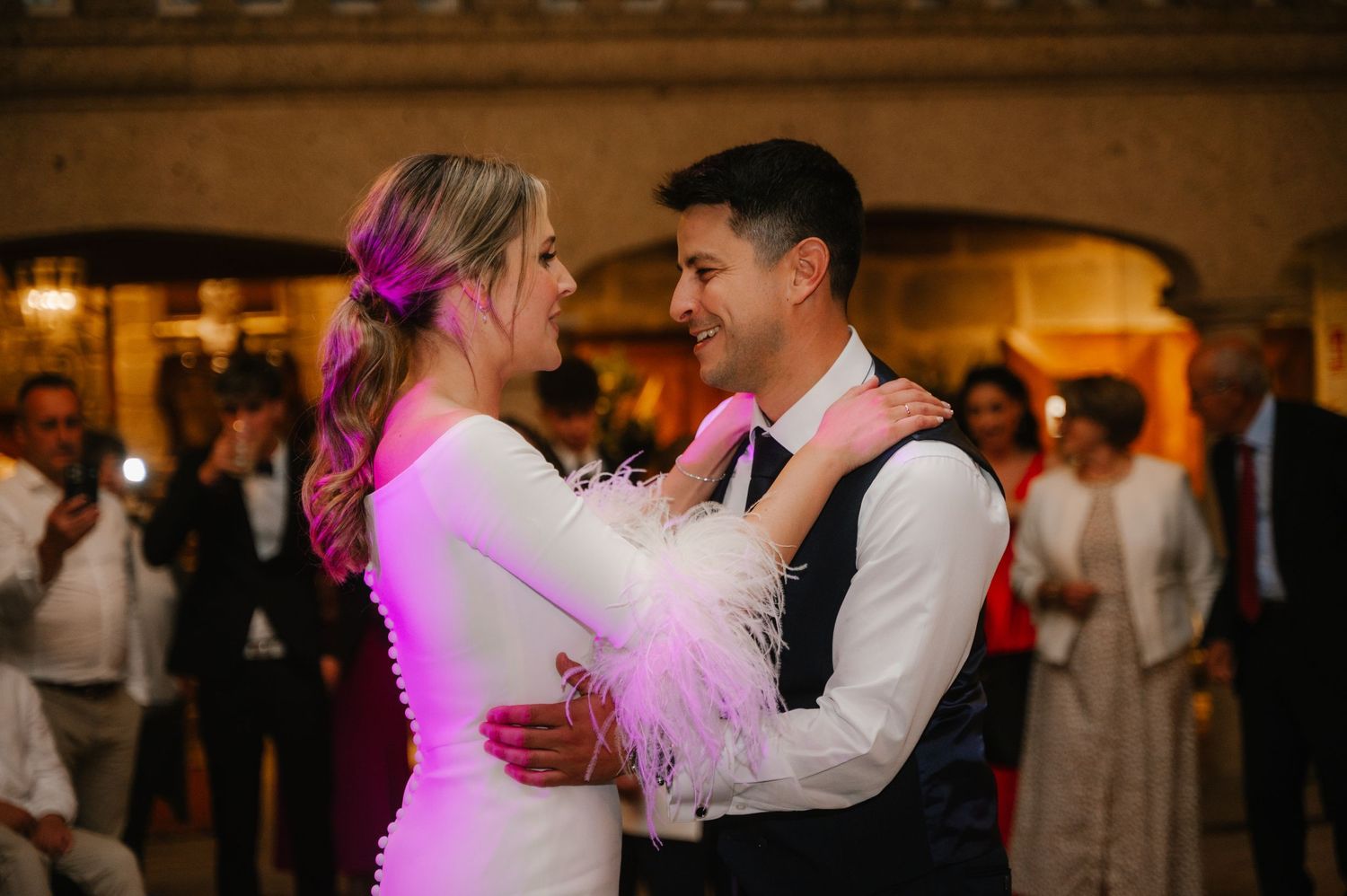 A couple shares a romantic dance at an elegant wedding reception with warm lighting and guests in the background.