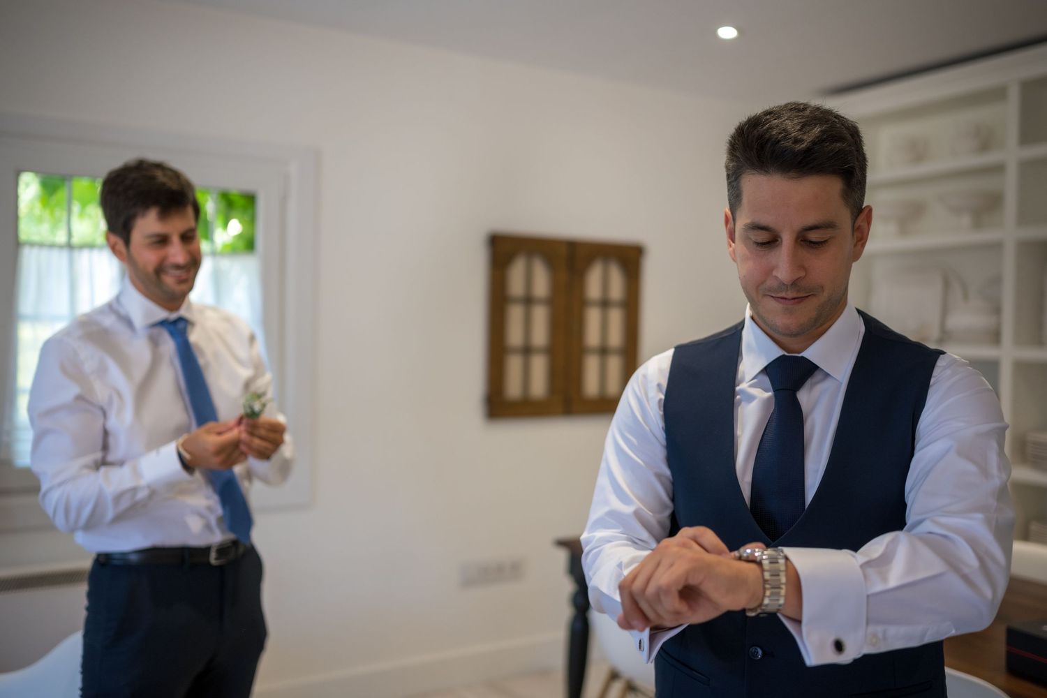 Groomsmen in dark suits and white shirts prepare for a wedding ceremony in a bright room.