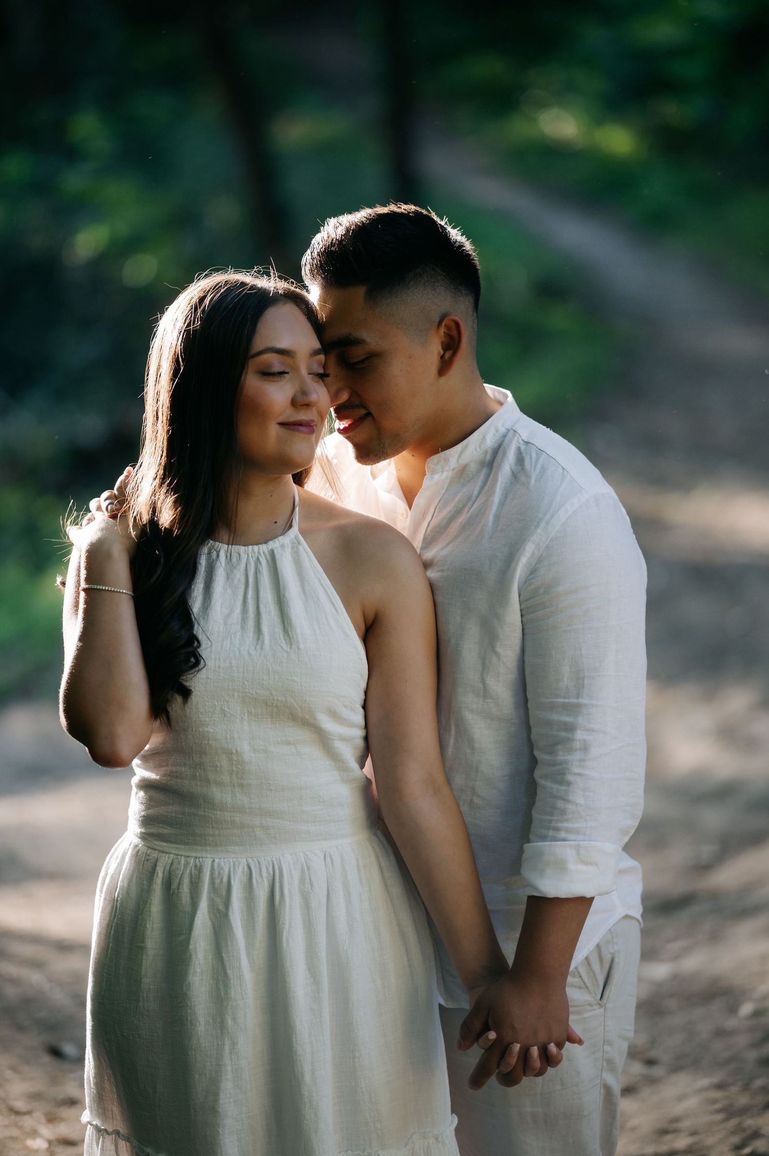 An intimate moment between a couple in white clothing captured in soft natural lighting on a woodland trail.