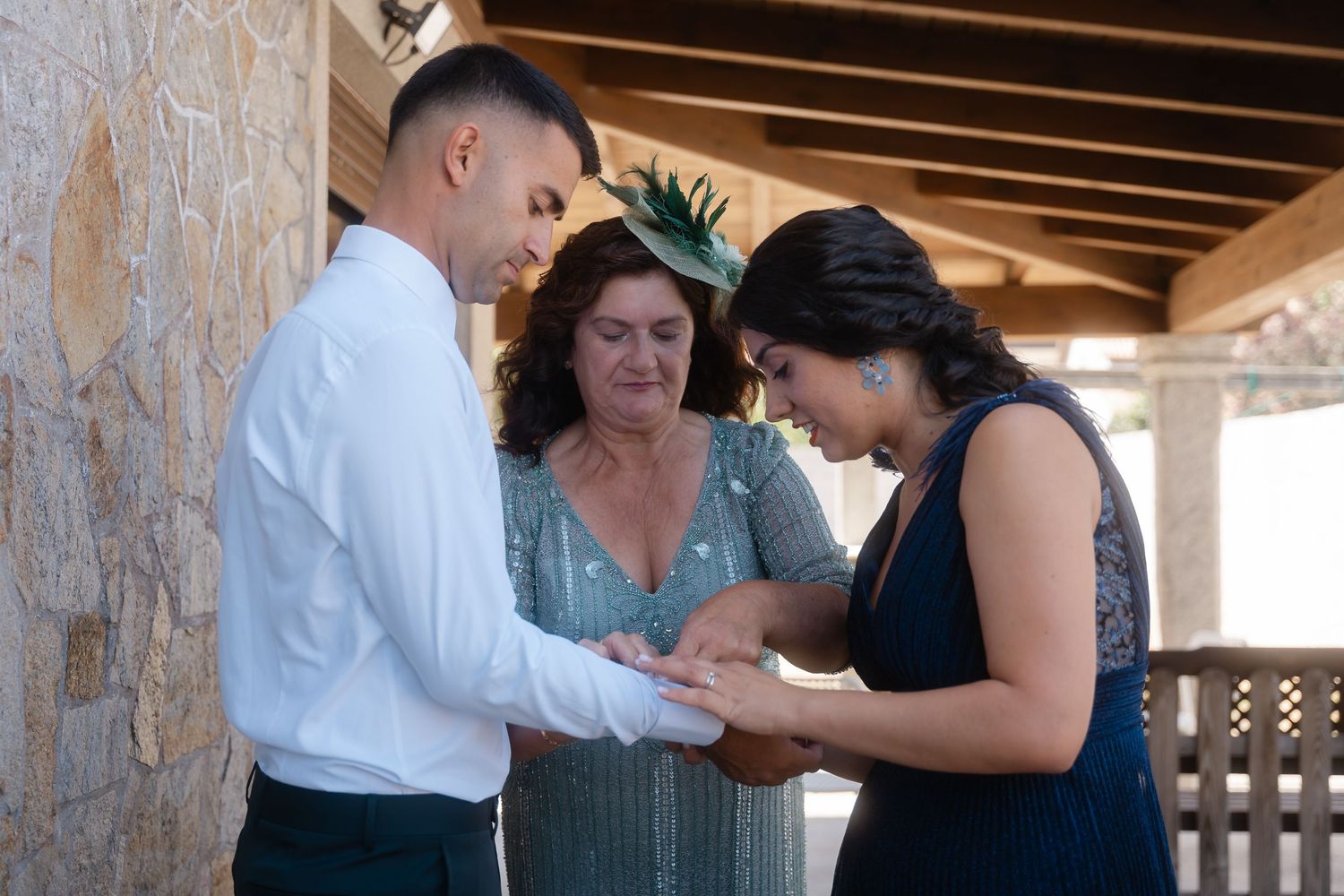 A couple exchanges rings during an outdoor ceremony with an officiant present in a covered porch setting.