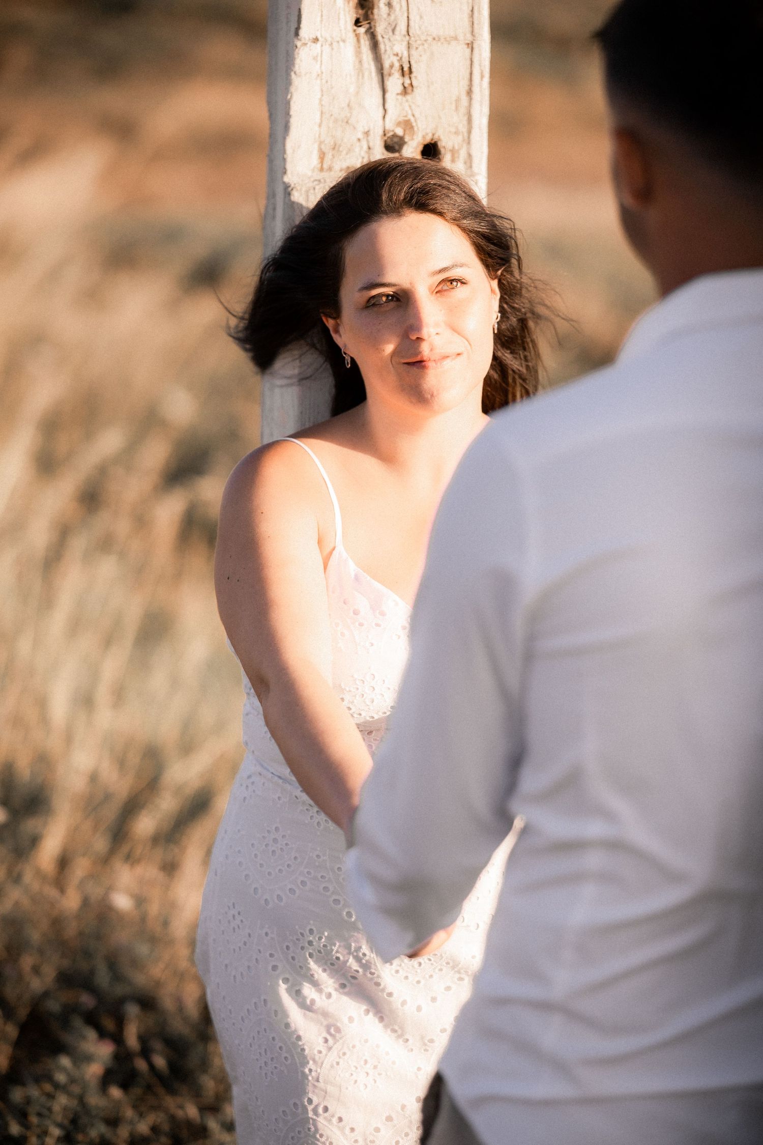 Intimate engagement photo of couple in white outfits against rustic backdrop during golden hour.