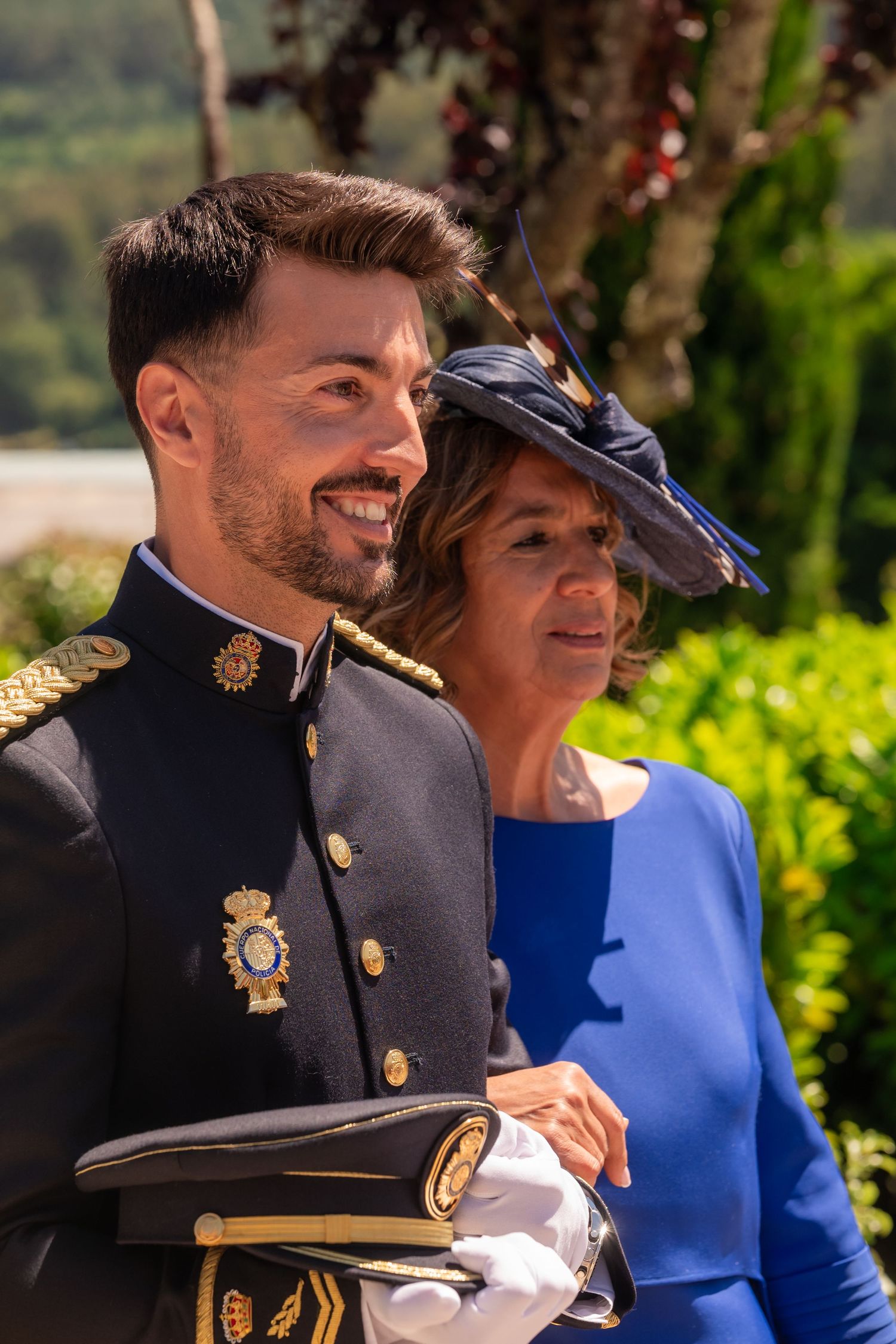 Military officer in dress uniform and woman in blue dress and hat smile together at outdoor celebration.